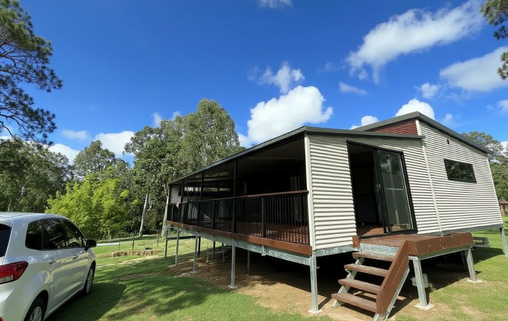 Cabin with a covered deck, on green grass, under a blue sky with clouds. White car is parked nearby — Steel Homes Australia in Branyan, QLD