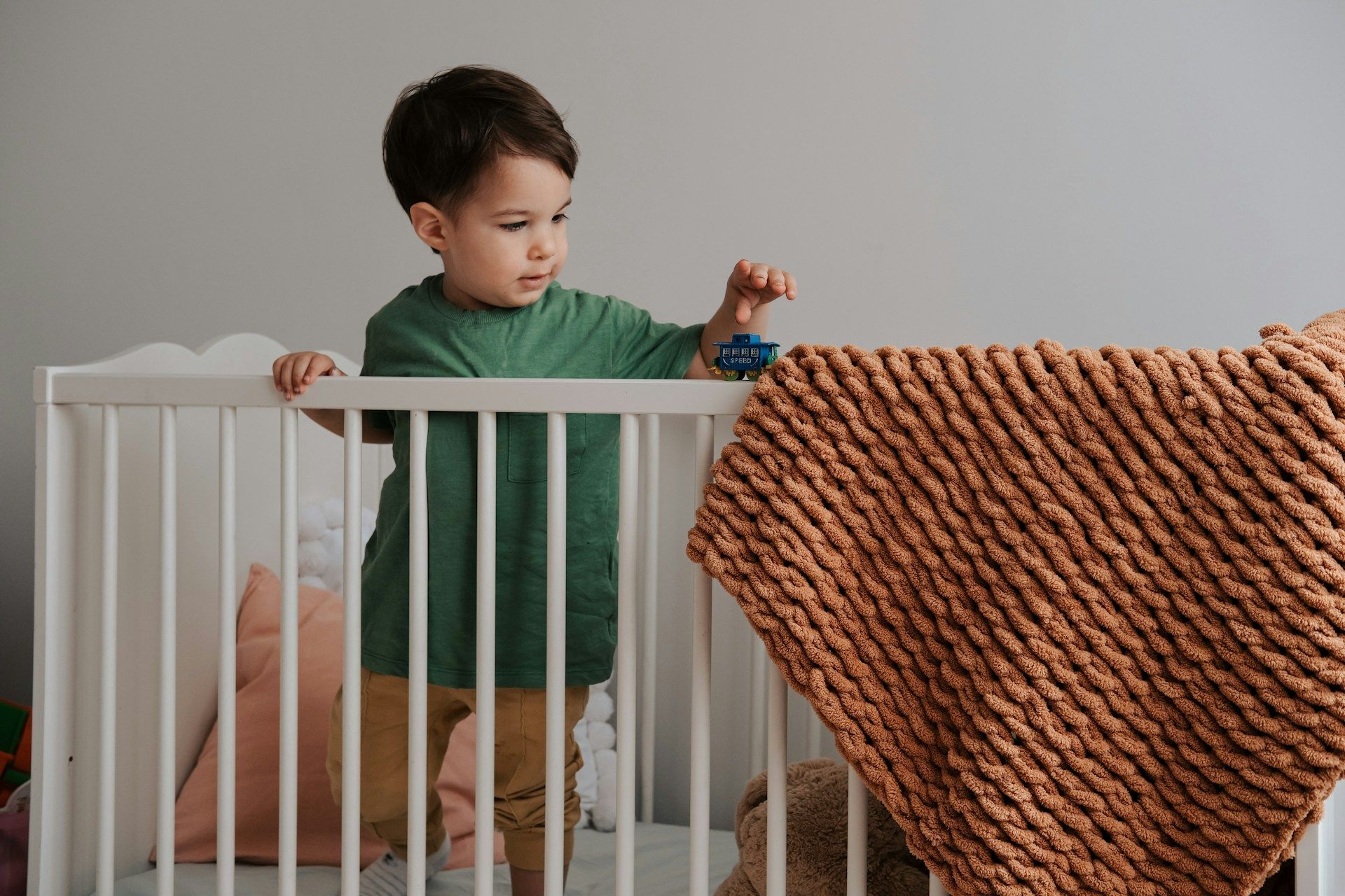 A young boy is standing in a crib holding a toy car.