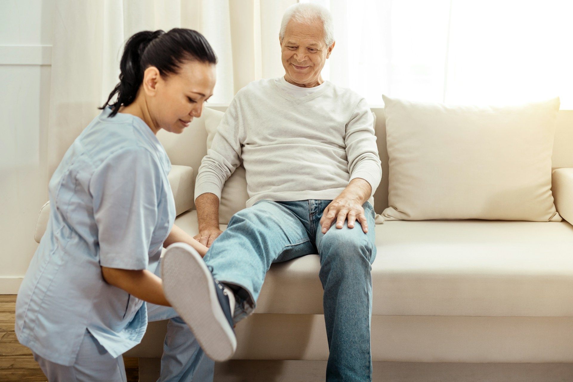 A nurse is helping an elderly man put on his shoes while sitting on a couch.