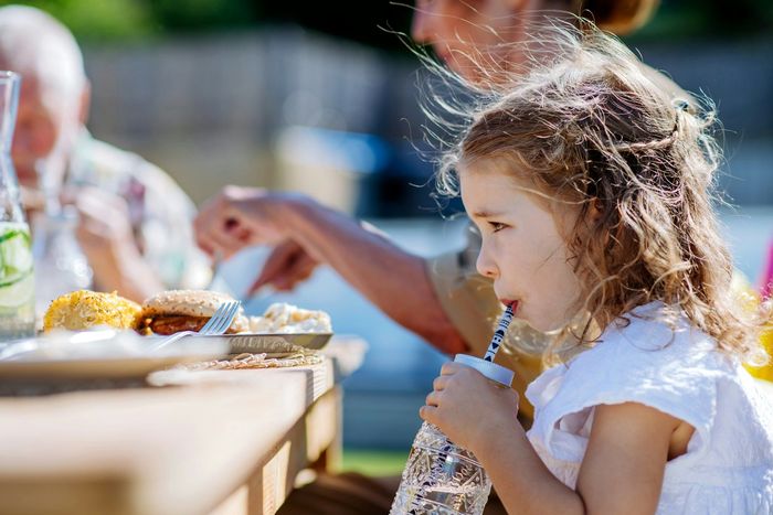 Girl drinks from a bottle with a straw at an outdoor table; other people and food are visible.