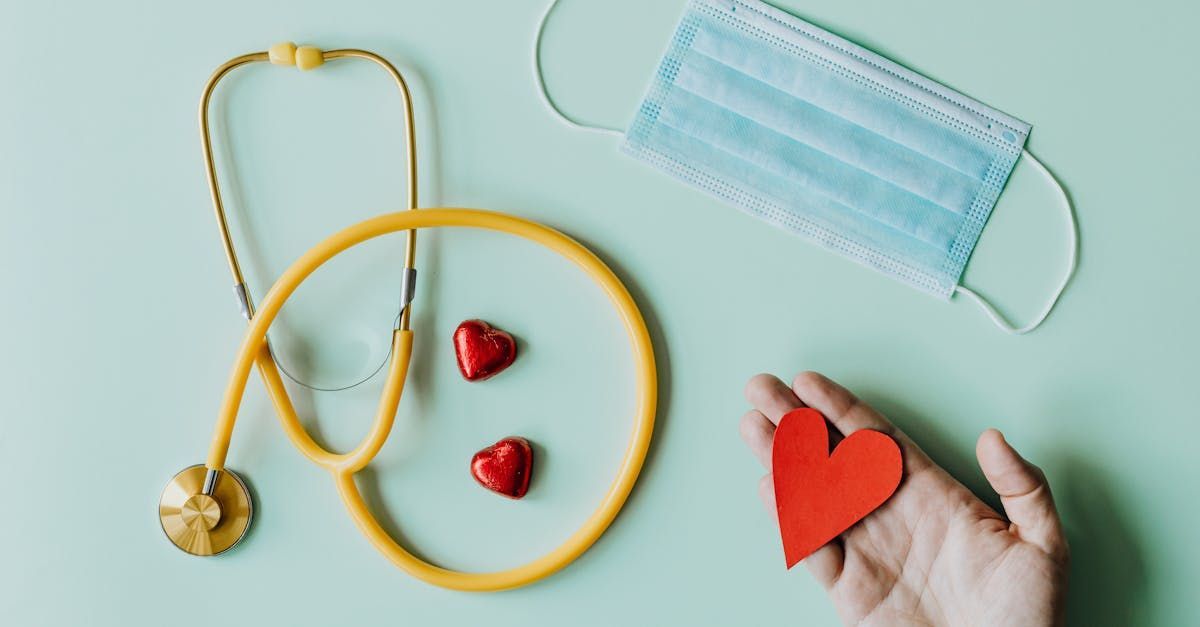 A person is holding a red heart in their hand next to a stethoscope and a mask.