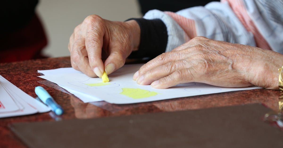 An elderly woman is sitting at a table drawing on a piece of paper.