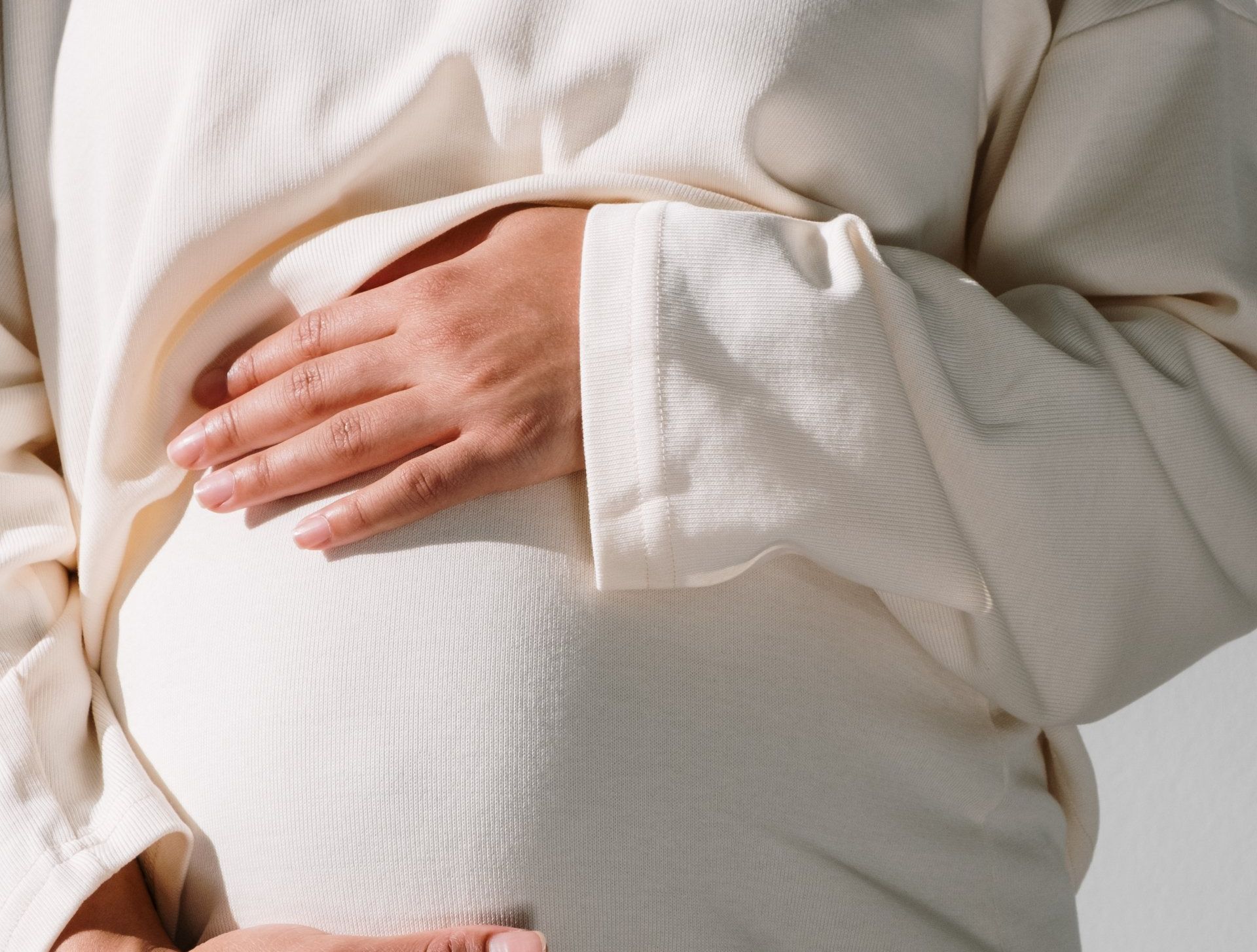 A pregnant woman in a white sweater is holding her belly.