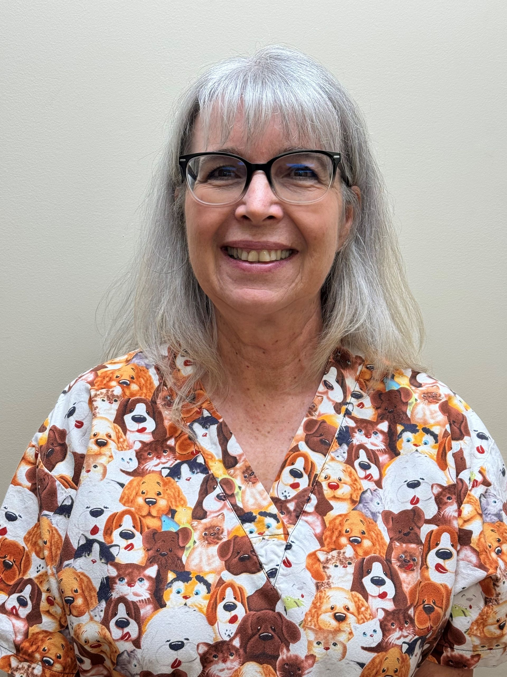 A woman wearing glasses and a scrub top with dogs on it is smiling for the camera.