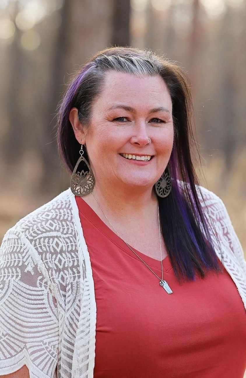 A woman with purple hair is wearing a red shirt and a white cardigan.