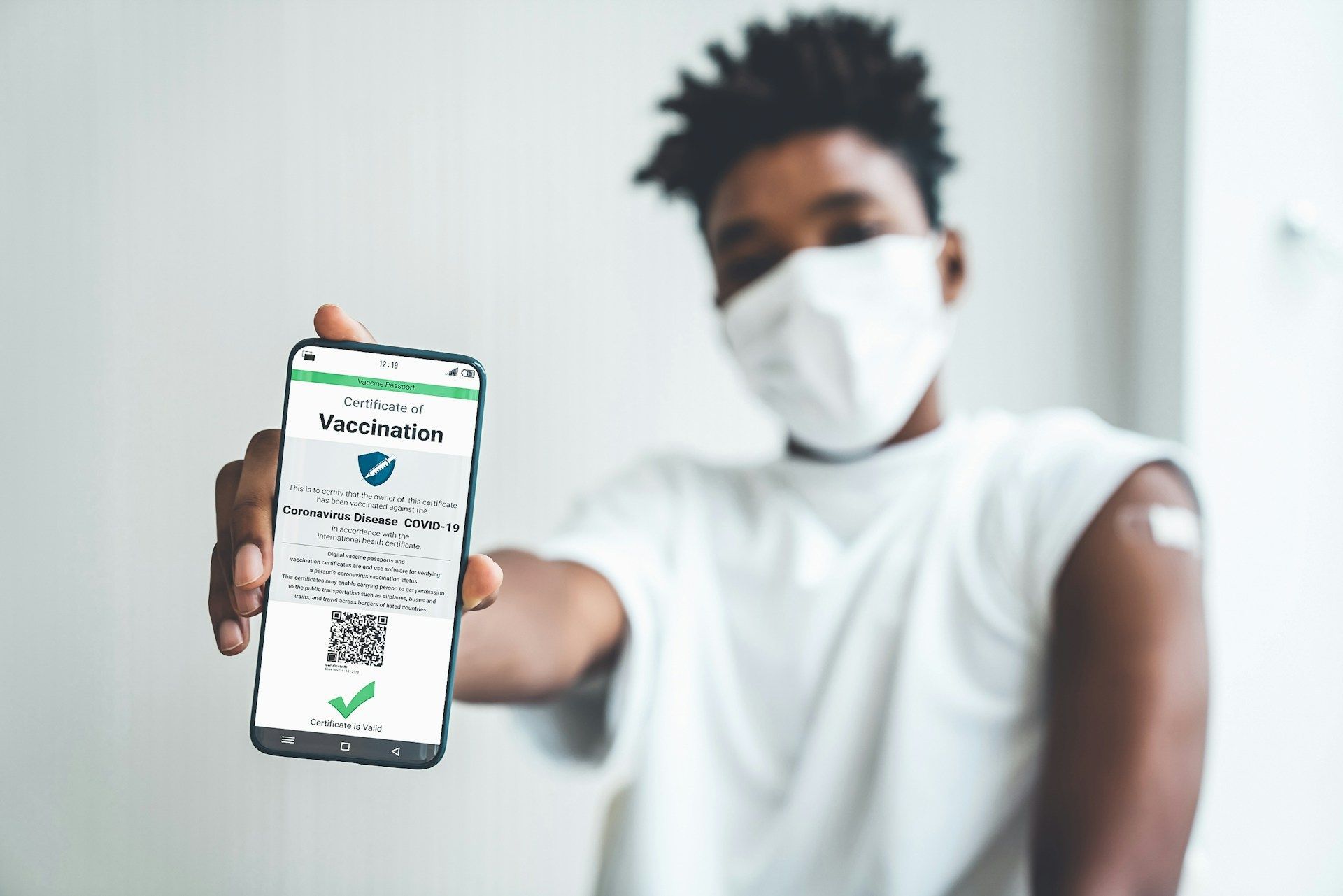 A man wearing a mask is holding a cell phone with a vaccination certificate on it.