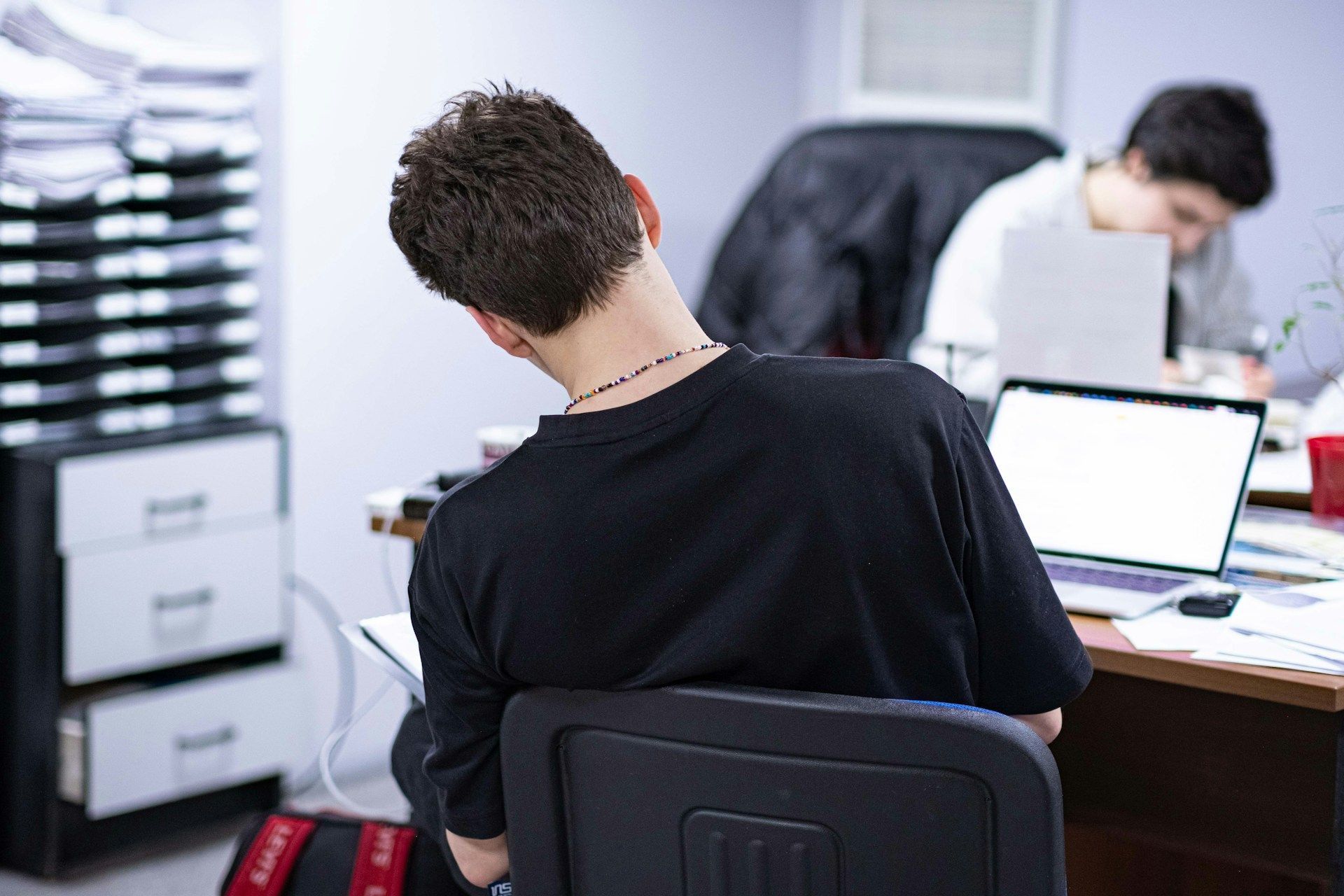 A man in a black shirt is sitting at a desk with a laptop.