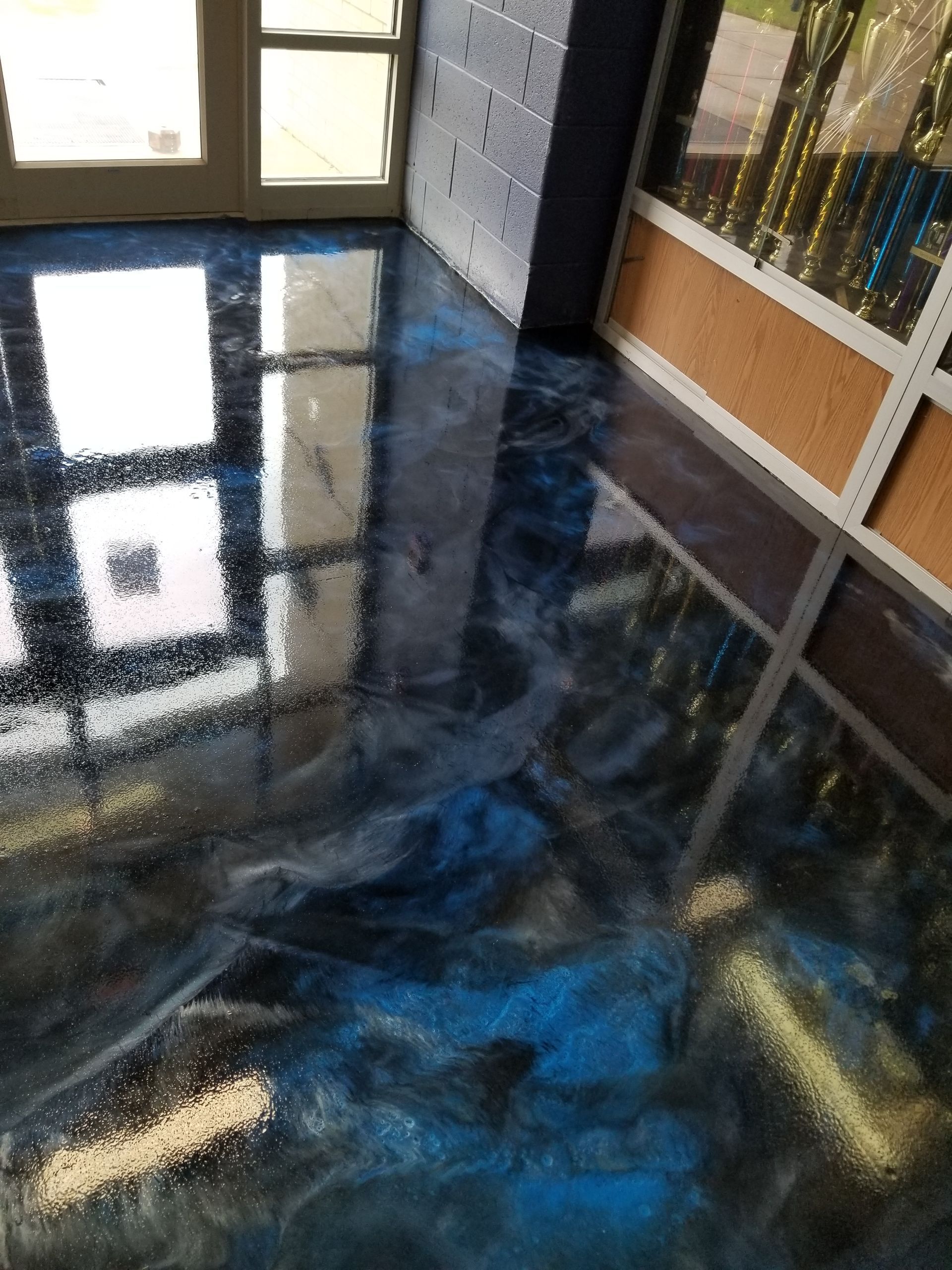 Shiny, blue and silver metallic epoxy flooring indoors, reflecting sunlight. A glass door and a wall of trophies are visible.