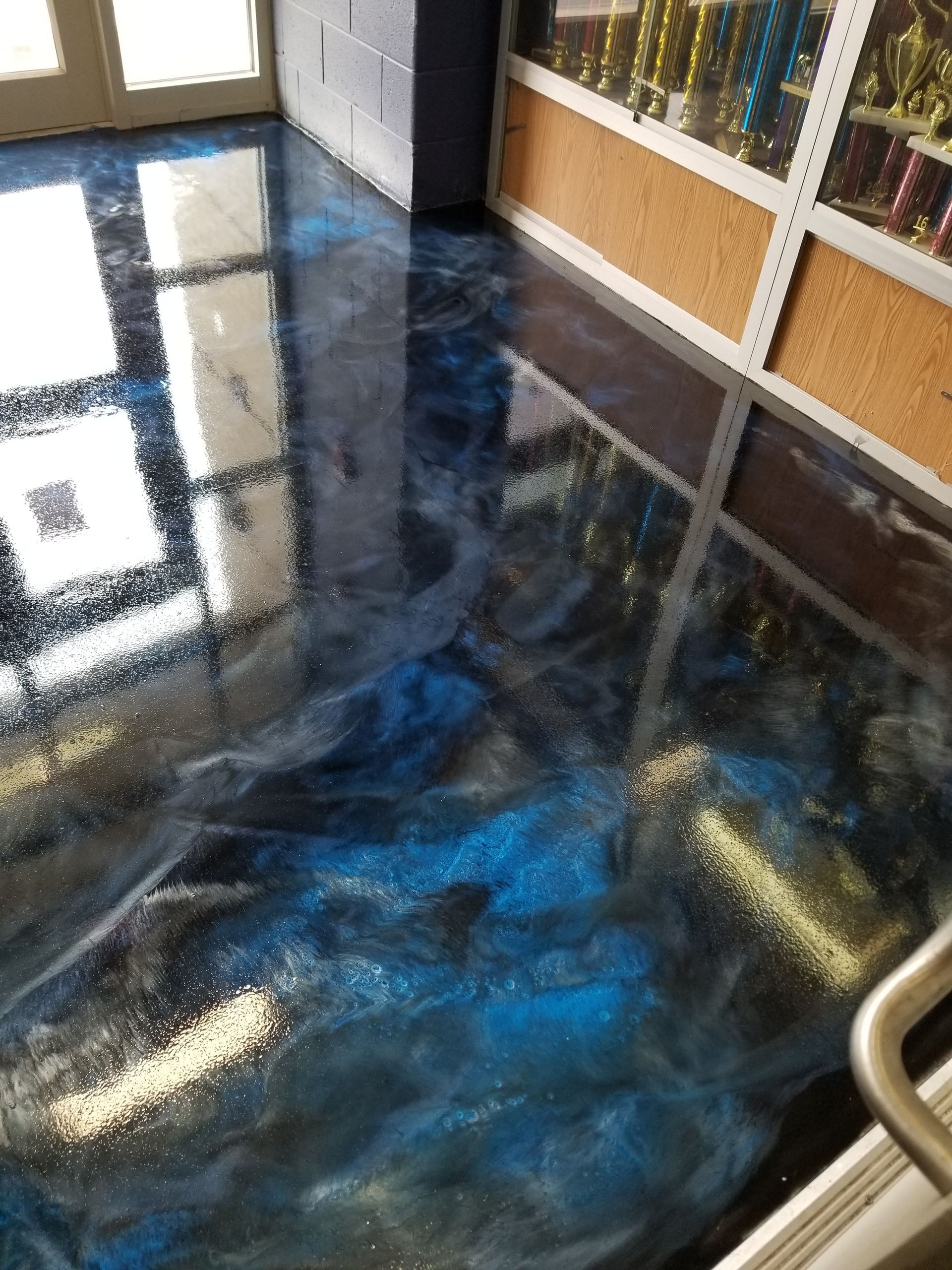 A glossy, epoxy-coated floor with blue, silver, and white swirls reflecting light. In a room with a window and trophy case.