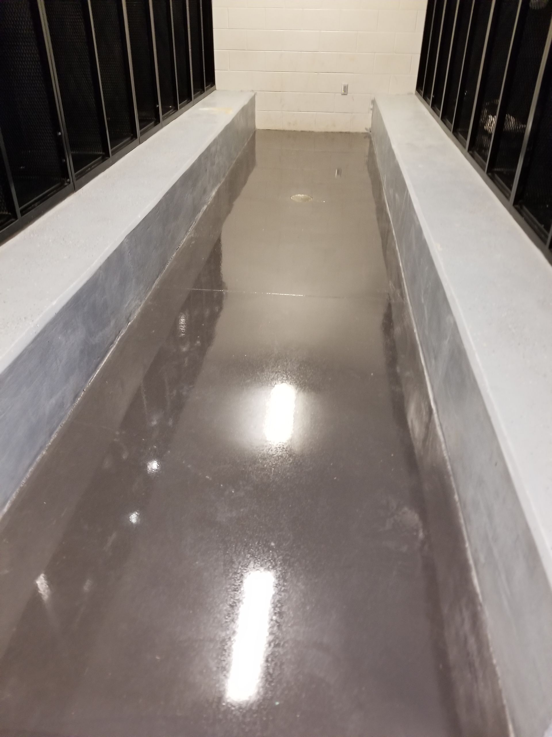 Narrow, gray-floored passageway with concrete borders. Metallic structures line both sides, and bright lights reflect off the wet-looking surface.