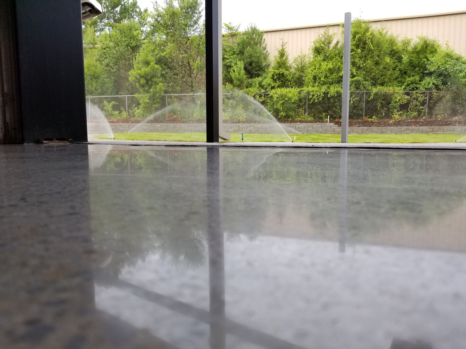 A polished concrete floor reflects an outdoor scene with sprinklers watering a green lawn, framed by a dark column and trees.