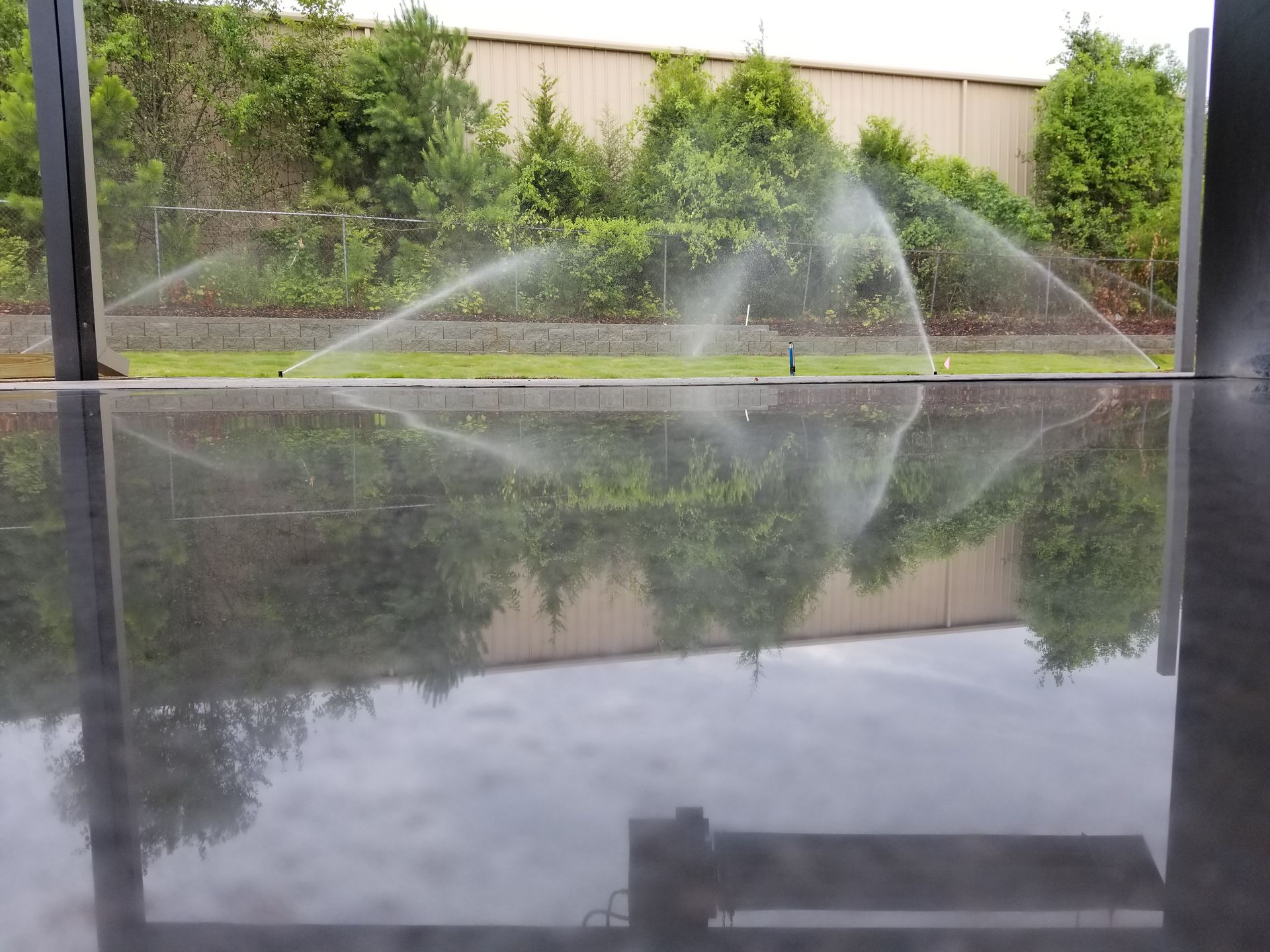Sprinklers watering a lawn, with water reflecting on a dark, wet surface in the foreground. Green plants and a tan building are in the background.