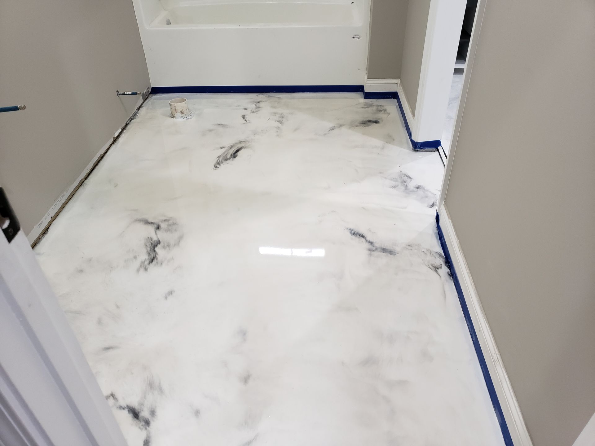 A bathroom with a white and black marbled epoxy floor. Blue tape lines the edges. Gray walls and a white bathtub are visible.
