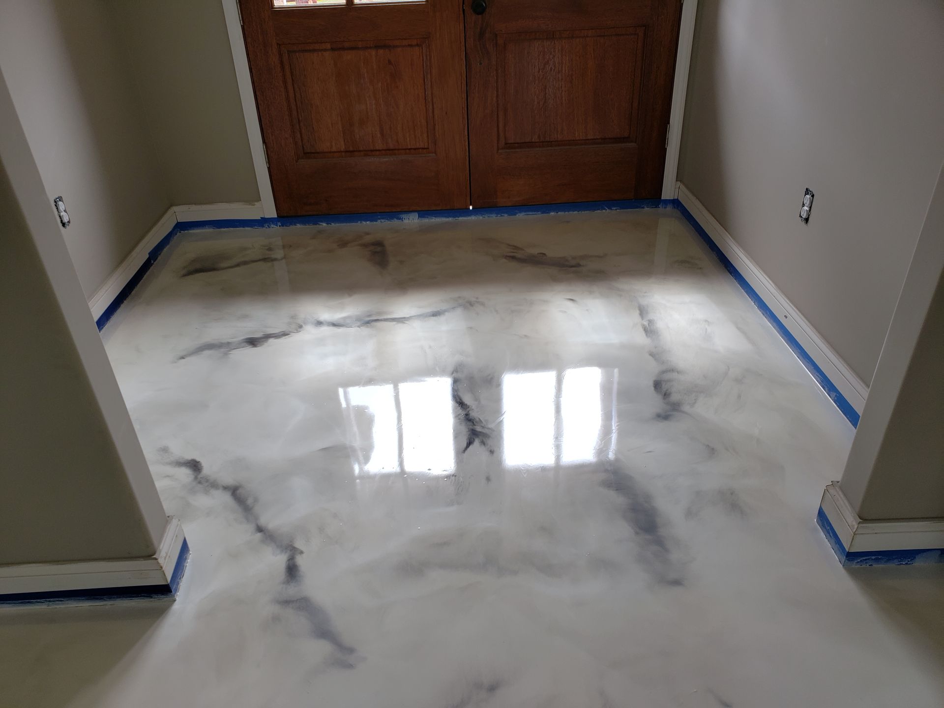 Metallic epoxy floor with marble effect in an entryway, reflecting light from a window. Blue tape protects the edges.