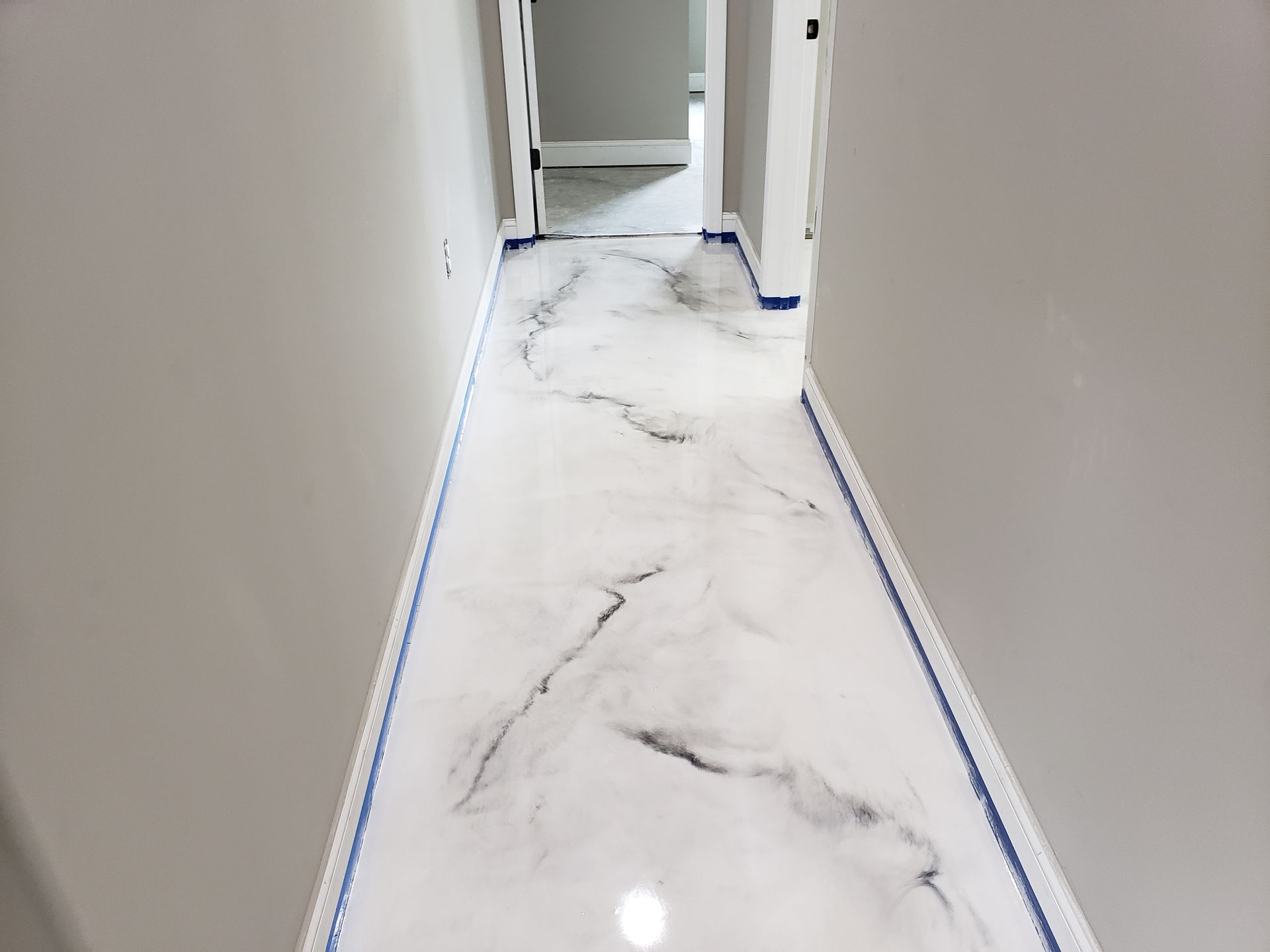 Hallway with shiny white and black epoxy floor, gray walls, and open doorways. Blue tape lines the floor edges.