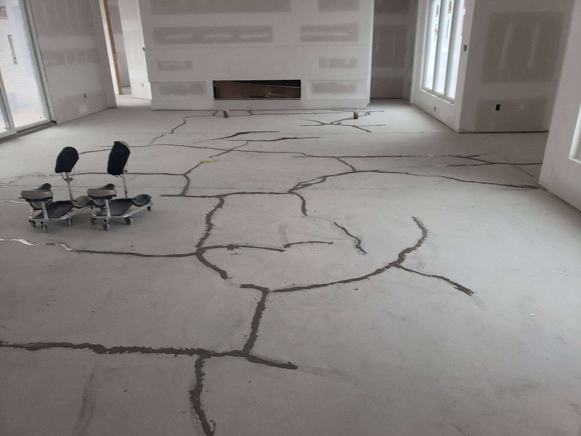 Cracked concrete floor in a room under construction. Grey cracks are filled with a dark material; two rolling chairs sit on the left.