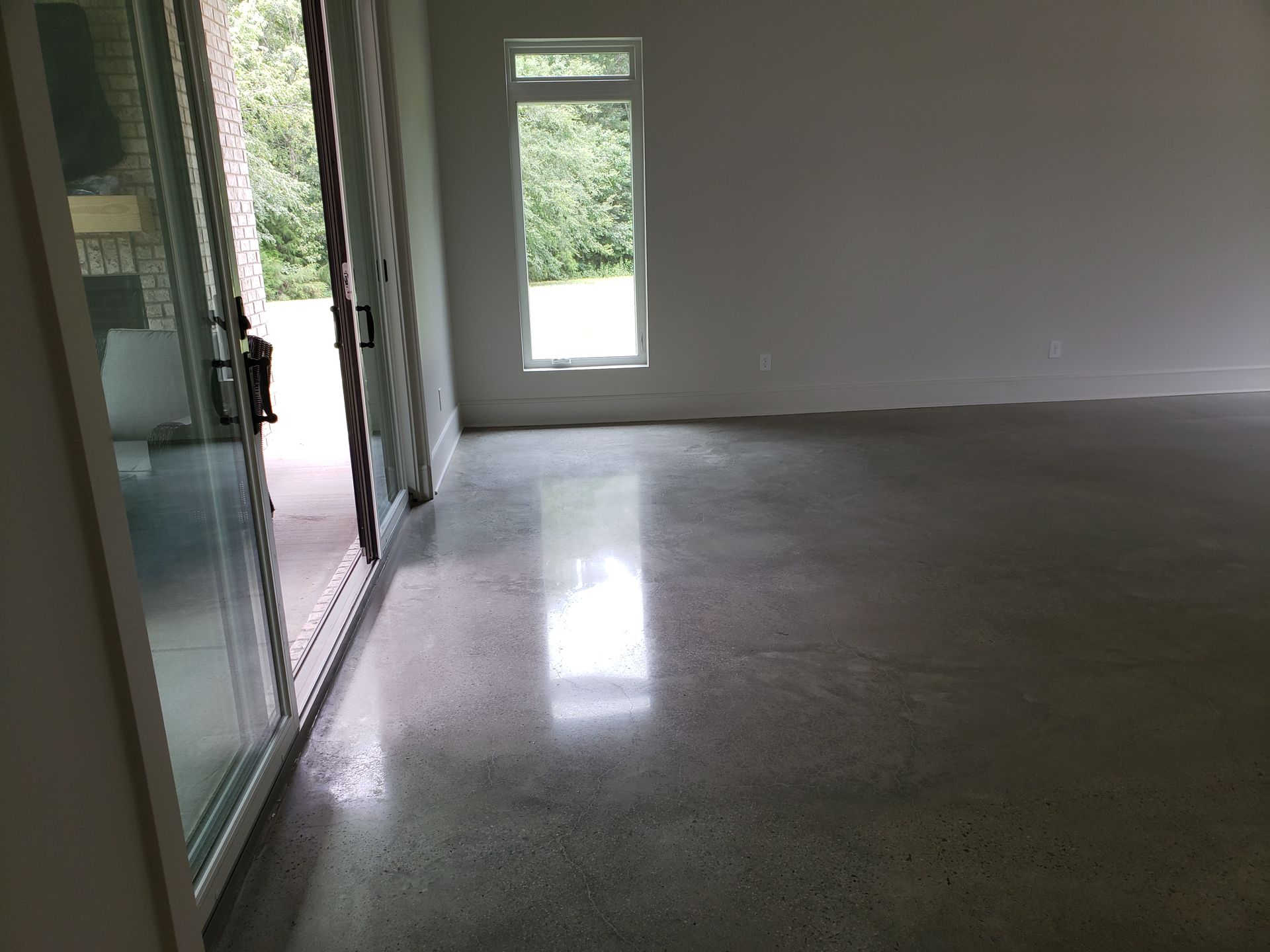 Polished concrete floor in a modern room. Sliding glass doors and a tall window provide outside views; walls are white.
