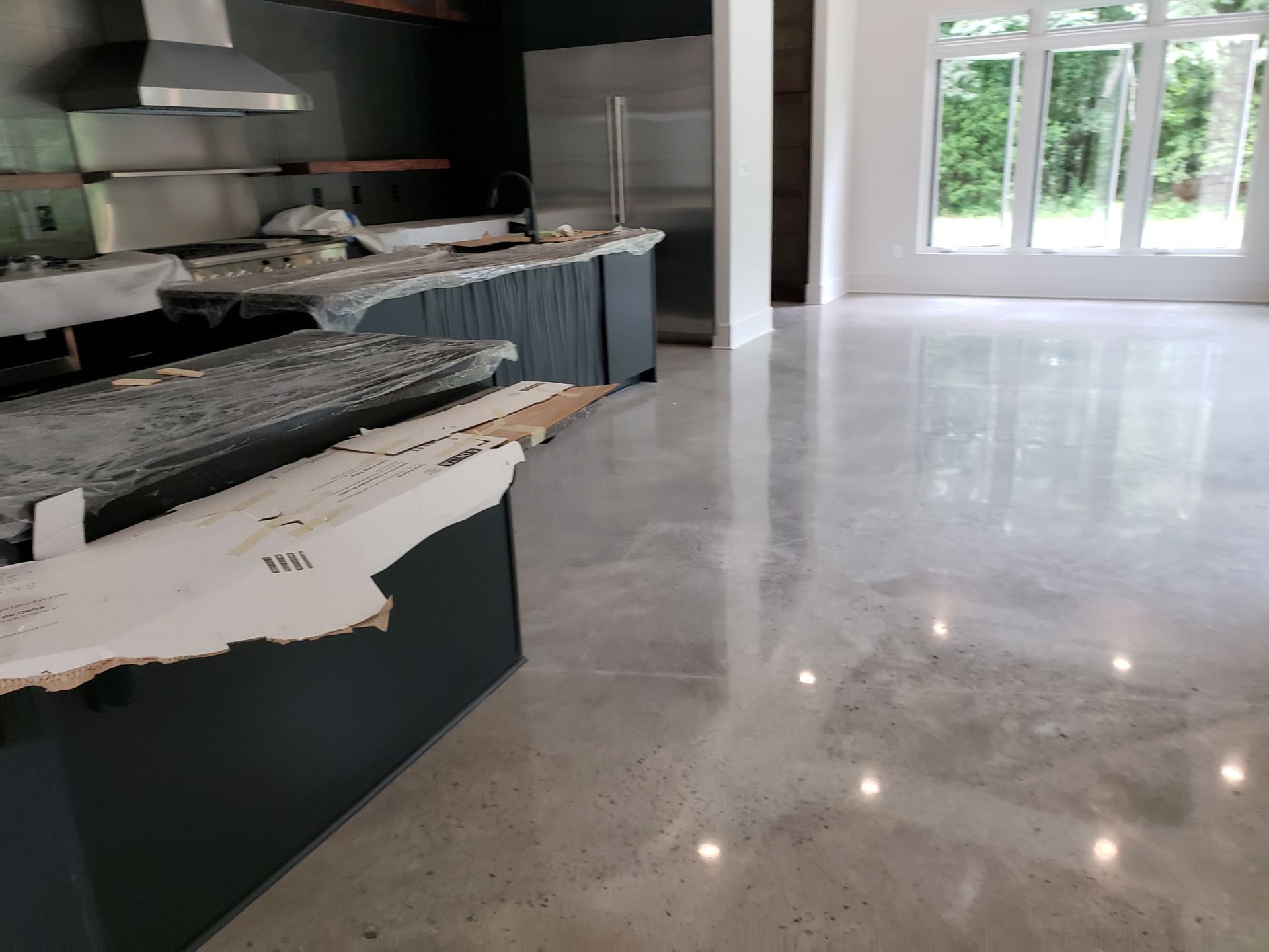 Kitchen with polished concrete floor, dark gray cabinets, and a stainless steel range hood. Large windows with a view of trees.