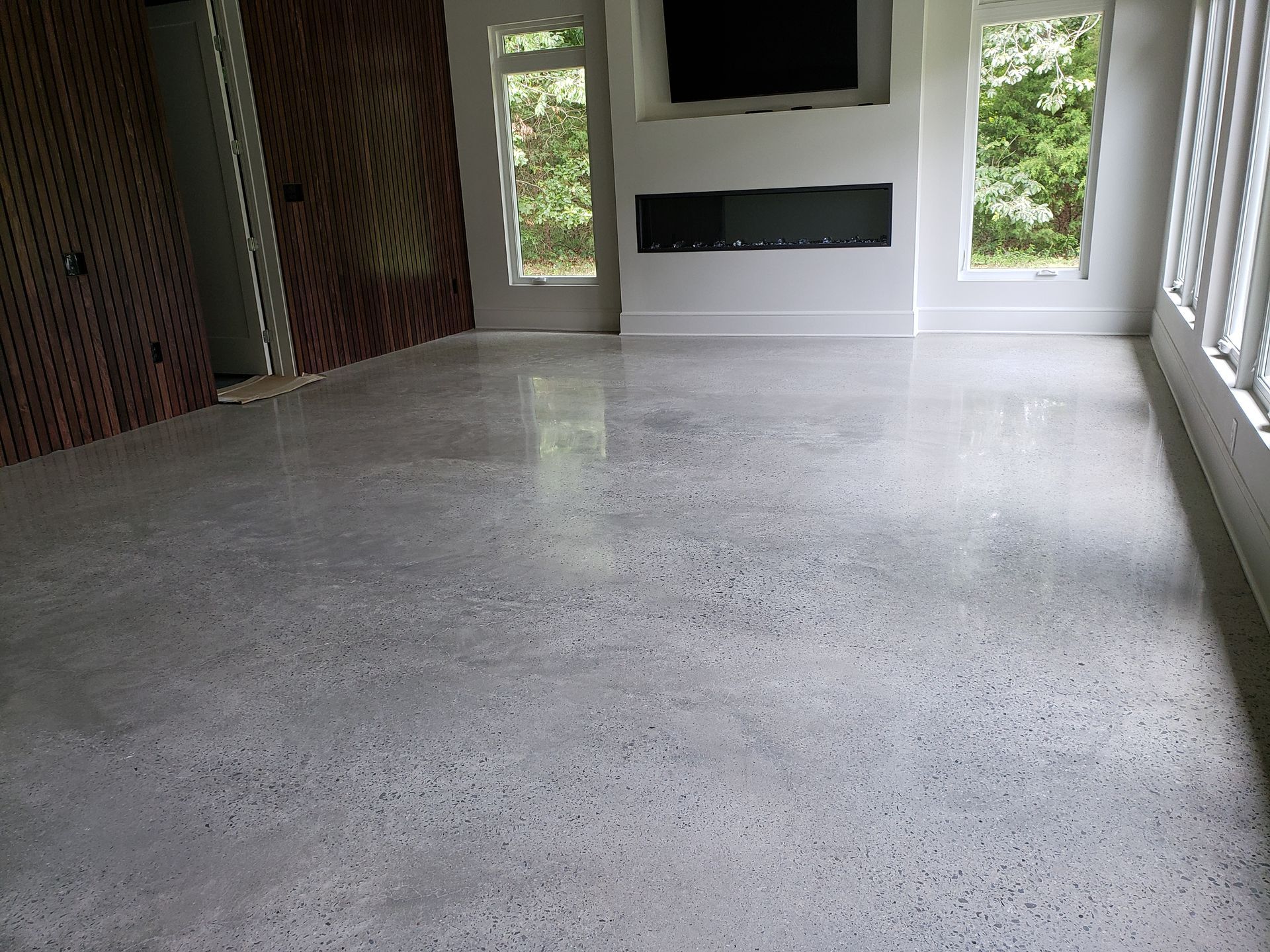Polished concrete floor in a modern room with a fireplace, windows, and a TV. Reflective gray floor with light streaming in.
