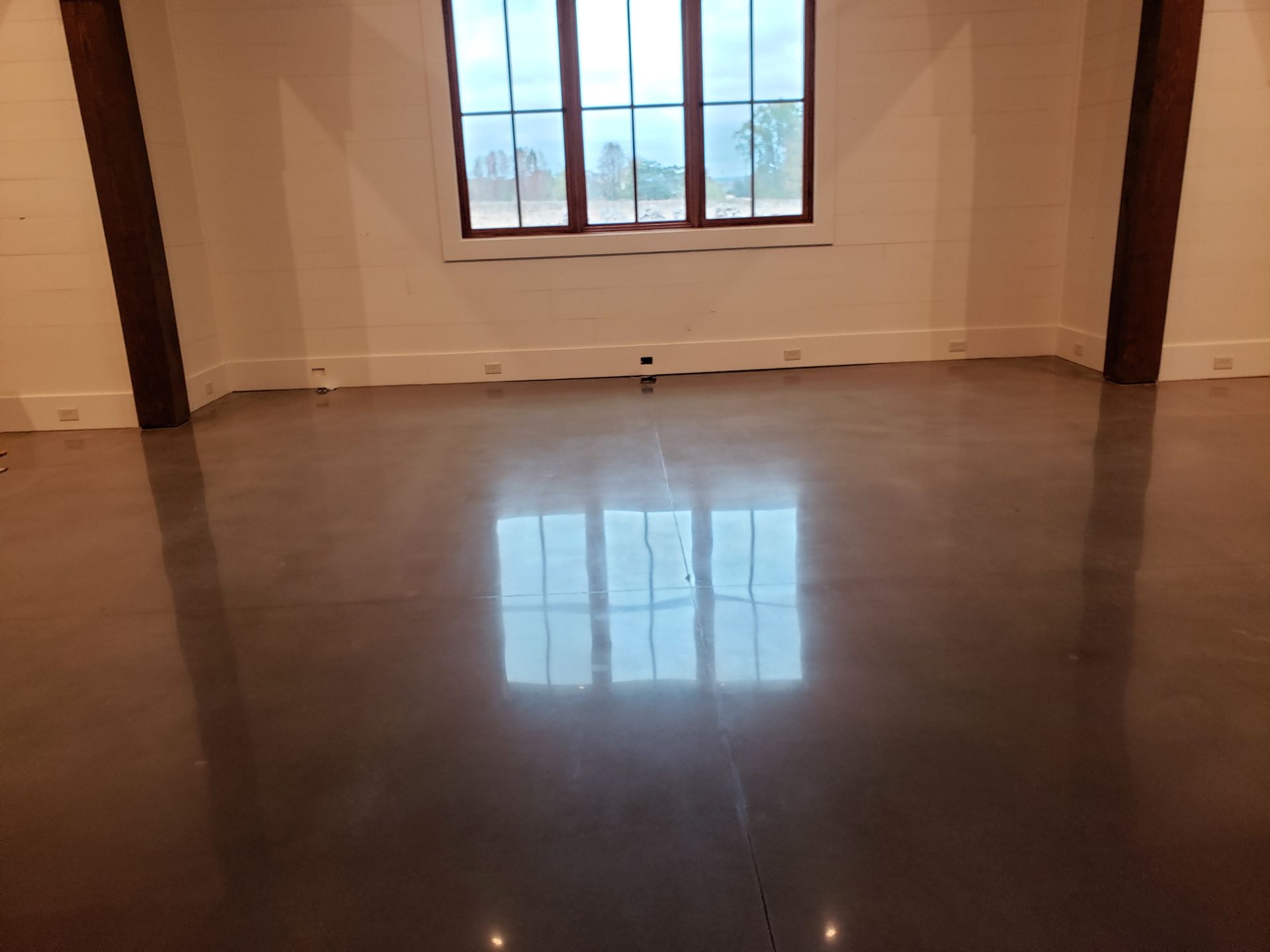 A polished concrete floor reflects a window, showing a bright, empty room with wooden beams and a white wall.