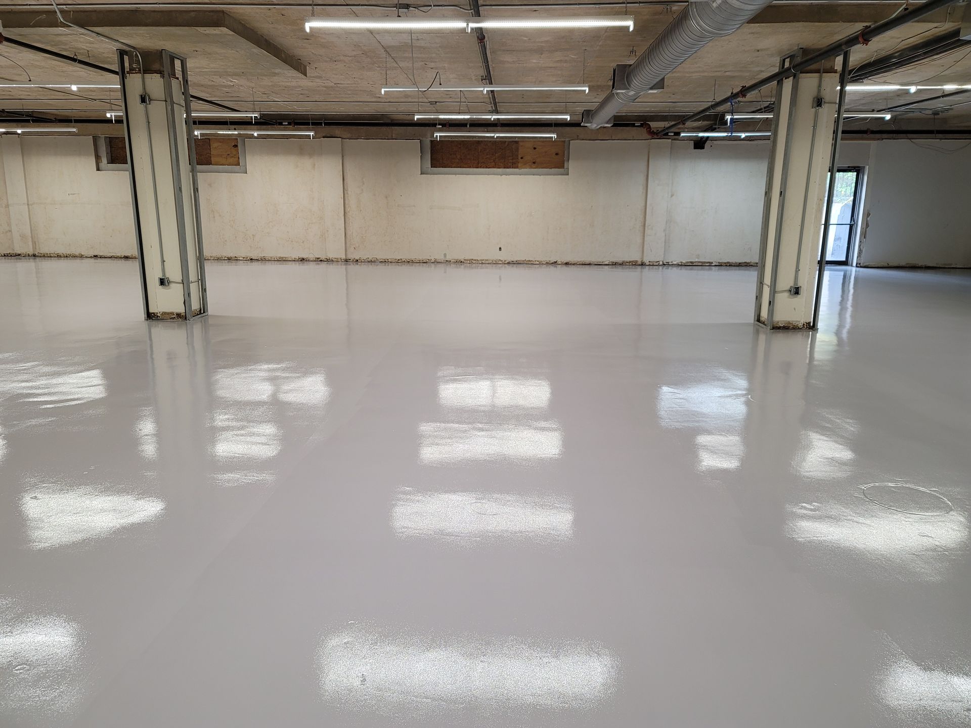 A wide shot of a freshly coated, light gray epoxy floor. The floor reflects the ceiling lights and concrete support pillars.