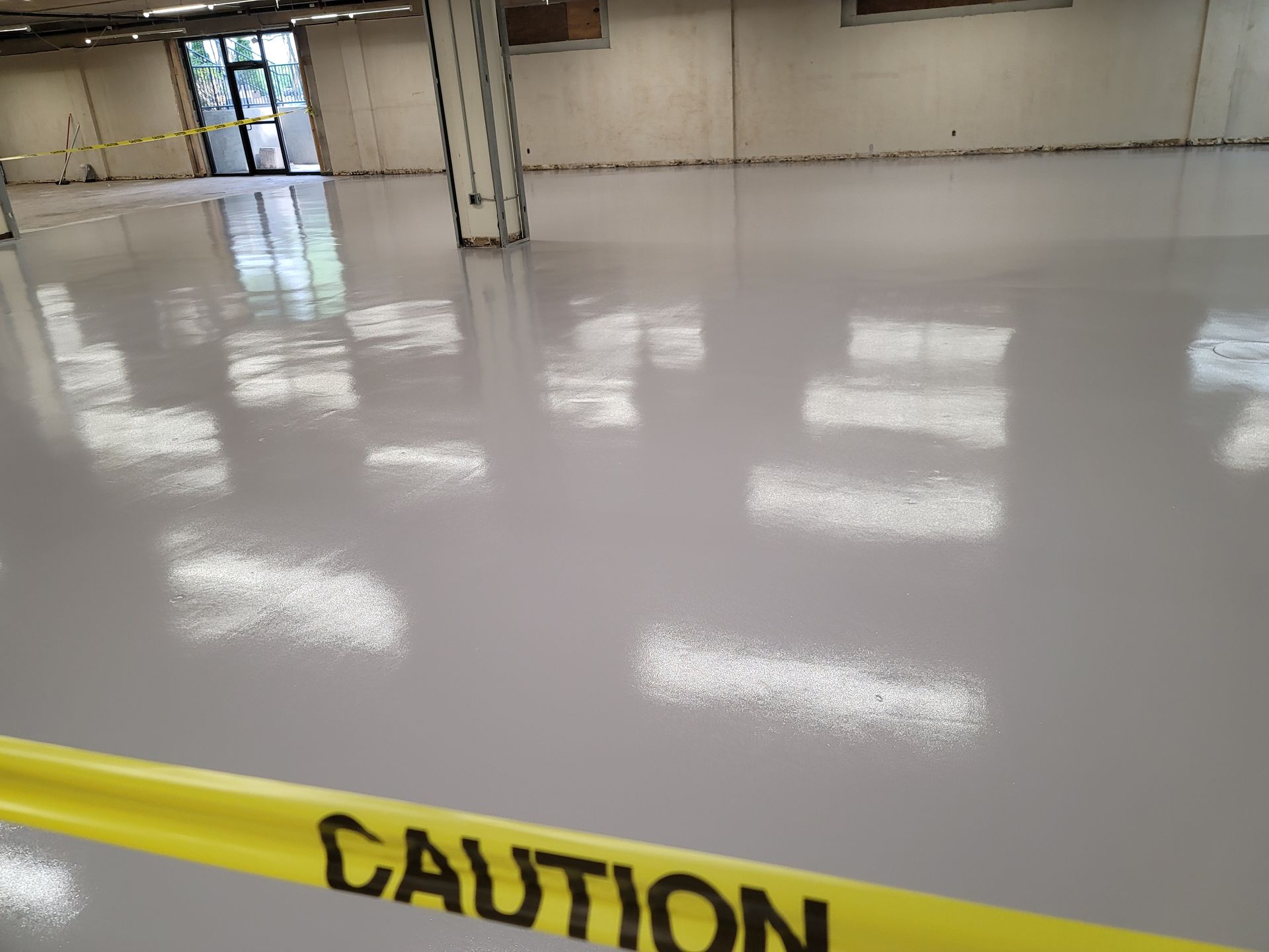 A freshly painted, light gray epoxy floor in a large, empty room with yellow caution tape in the foreground.
