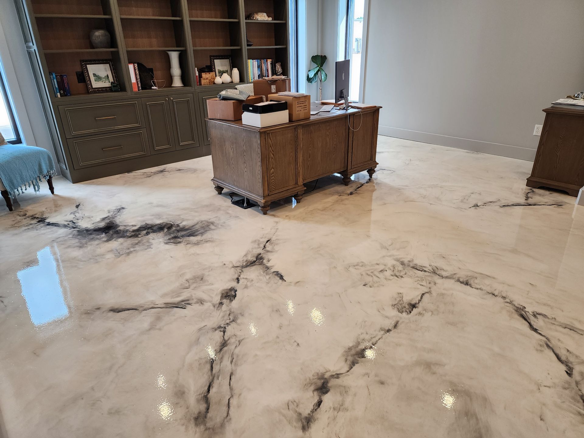 Room with marble-effect epoxy flooring, large wooden desk, bookshelves, and an additional cabinet. The floor reflects light, and the walls are gray.
