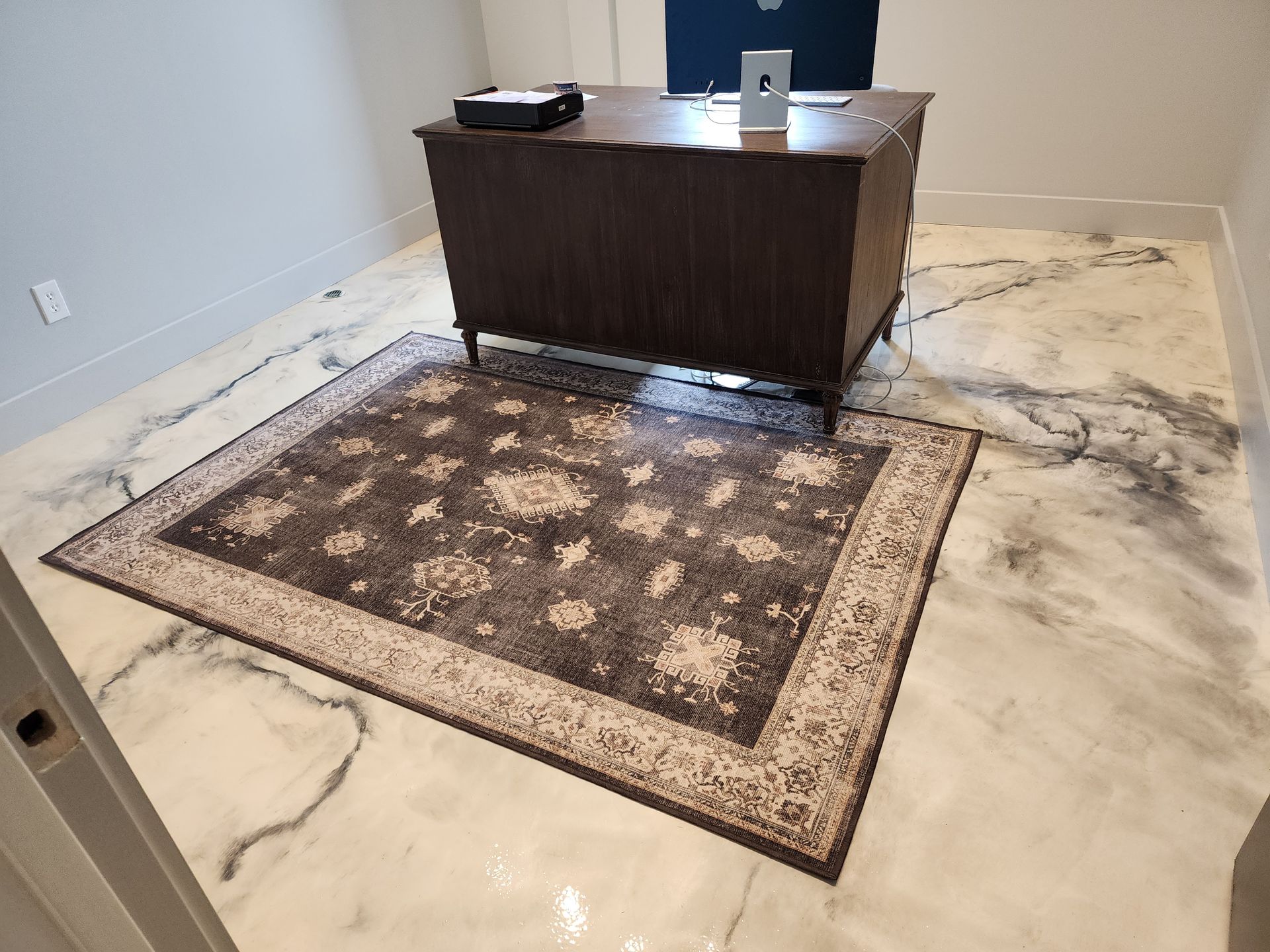 A dark wood desk sits on a patterned rug in a room with a metallic epoxy floor. A laptop and other objects are on the desk.