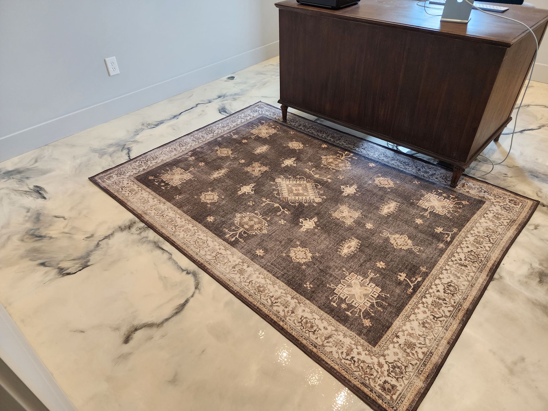 A dark brown rug with a beige border sits on a glossy, marbled floor in front of a dark wooden desk.