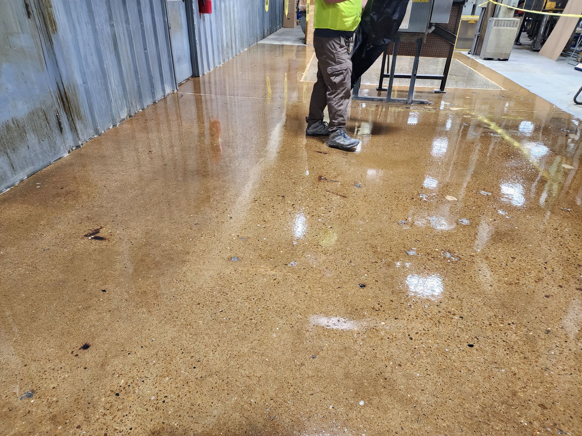 Person walking on a wet, polished concrete floor in a factory setting. The floor reflects the room's lights and features a brown hue.