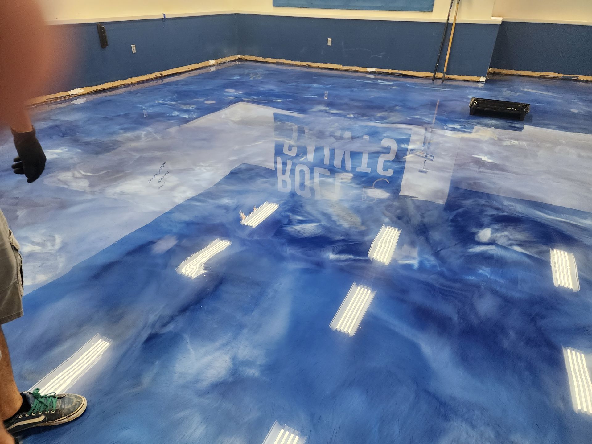 A person with black gloves stands in a gymnasium with a blue and white epoxy floor. The floor reflects the lights and writing on a wall.