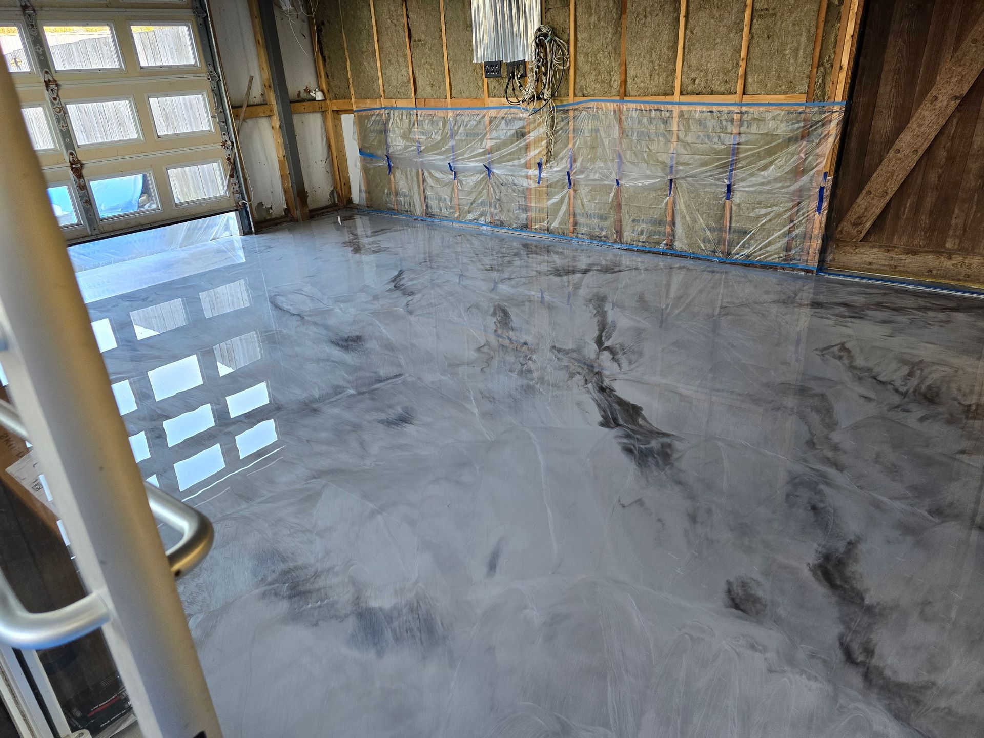 Metallic epoxy-coated garage floor reflecting sunlight and interior features. Walls show insulation and framing.