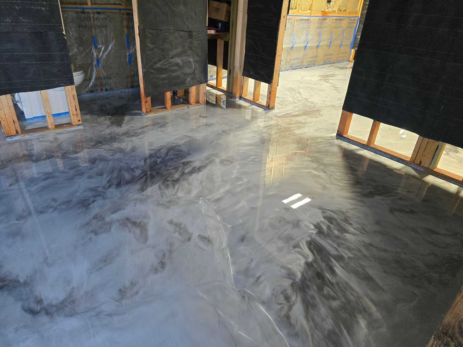 Metallic epoxy floor in a room under construction, featuring silver and dark grey swirling patterns. Wooden framing is visible.