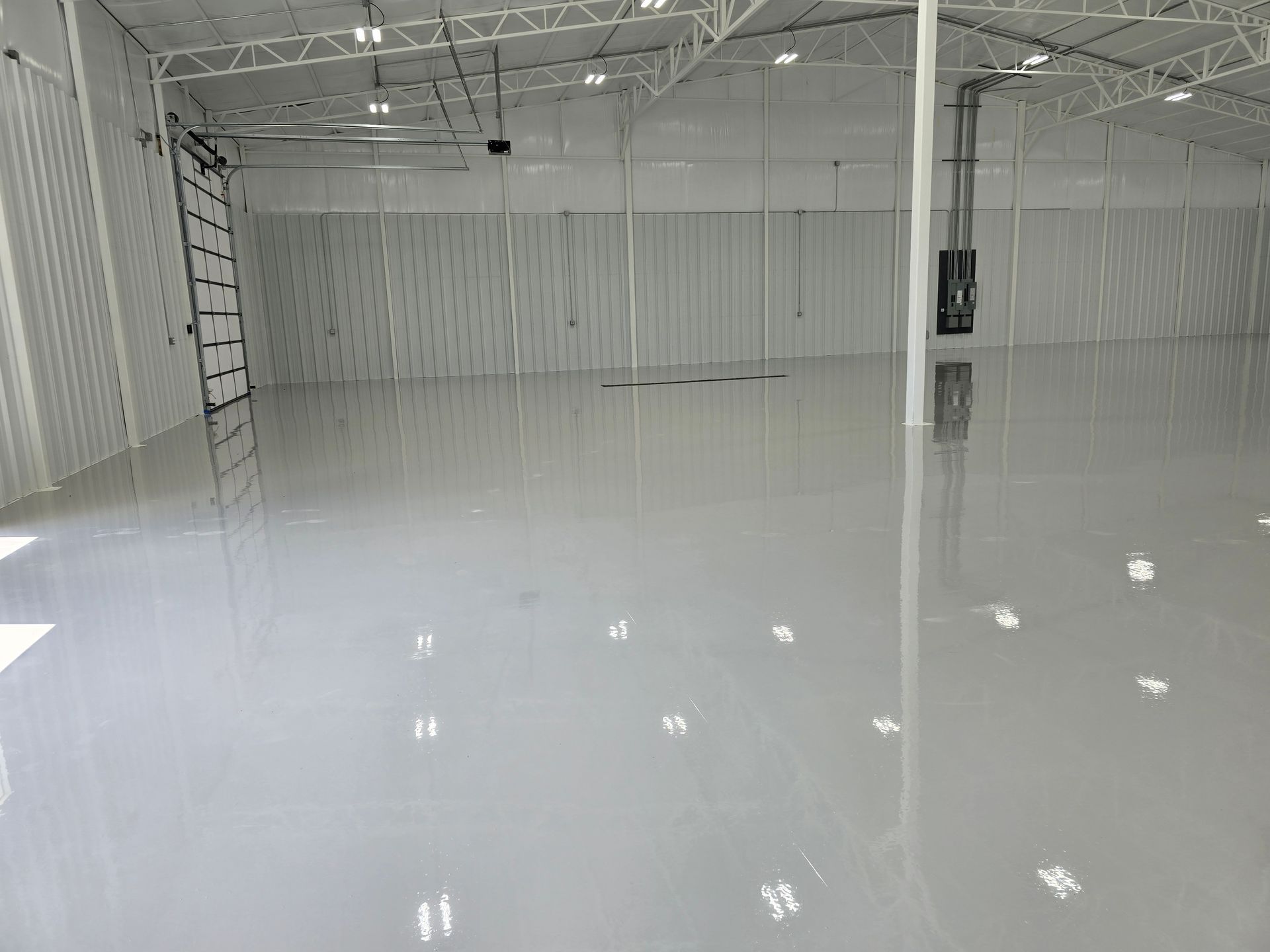 Empty, shiny grey-floored warehouse interior. White walls, high ceiling with lights and metal beams.