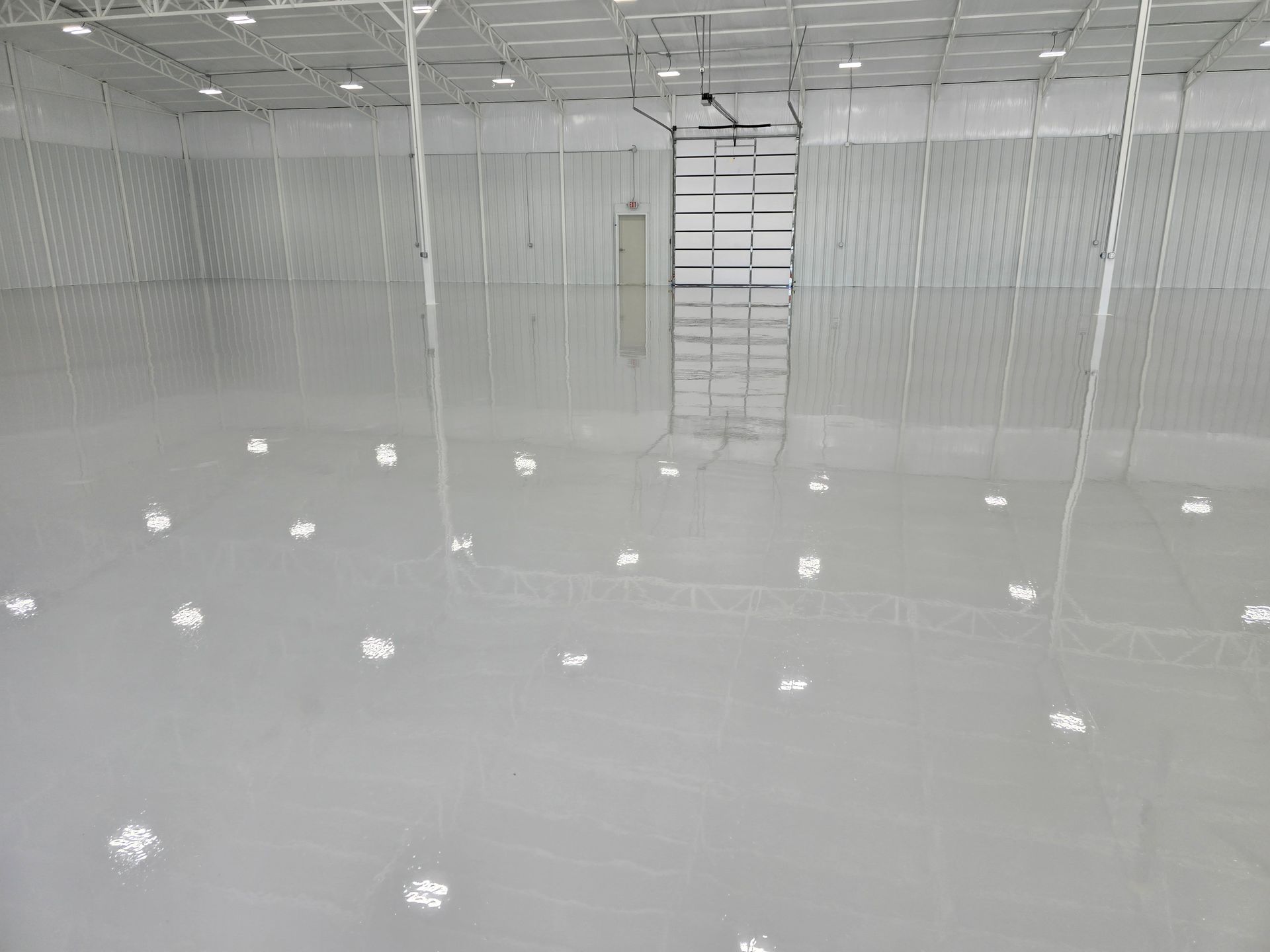 Interior of a large, empty warehouse with a shiny, gray floor reflecting overhead lights and a loading door at the end.