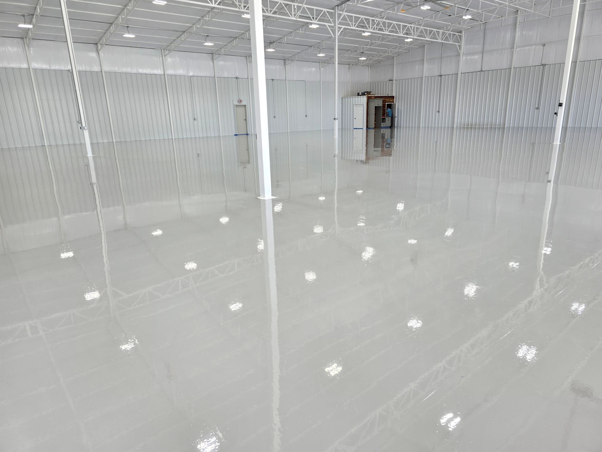 A large, empty warehouse interior with a shiny, reflective, light gray floor. The walls and ceiling are white.