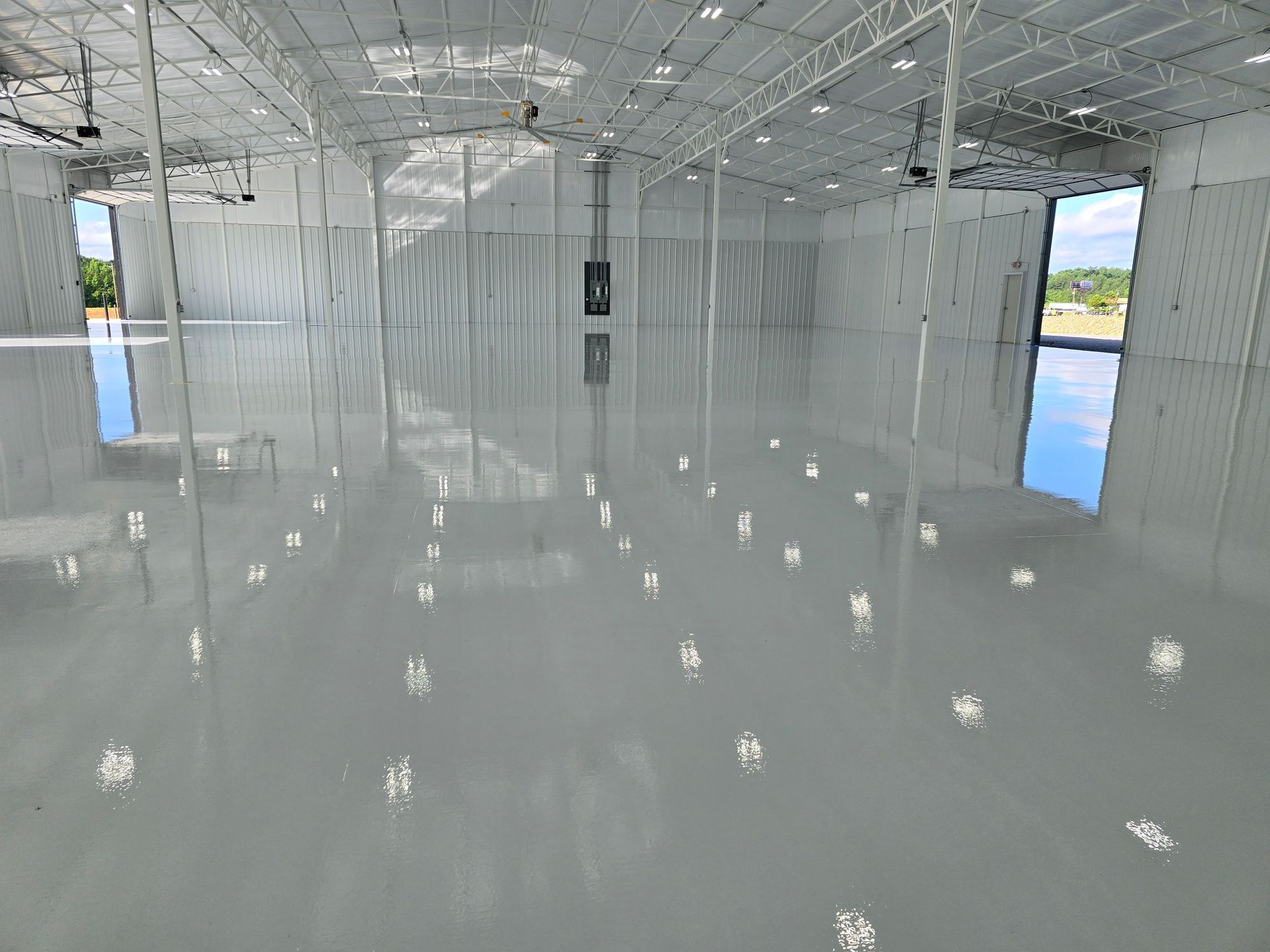 Spacious, empty warehouse interior with a shiny, gray epoxy floor. Reflections of the sky and structure are visible in the floor.