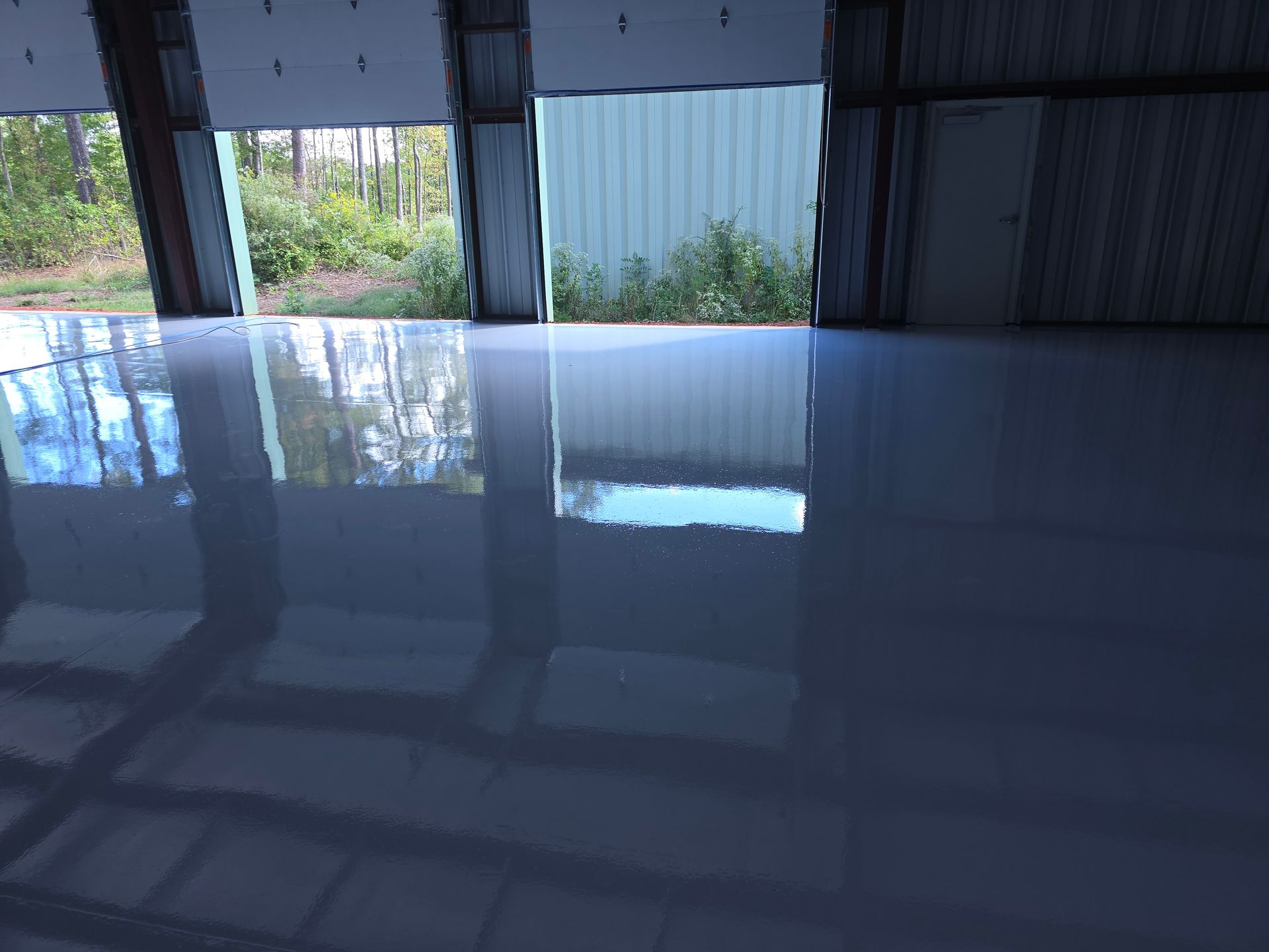 Glossy gray floor reflects a warehouse interior with open doors, showing a glimpse of trees outside.