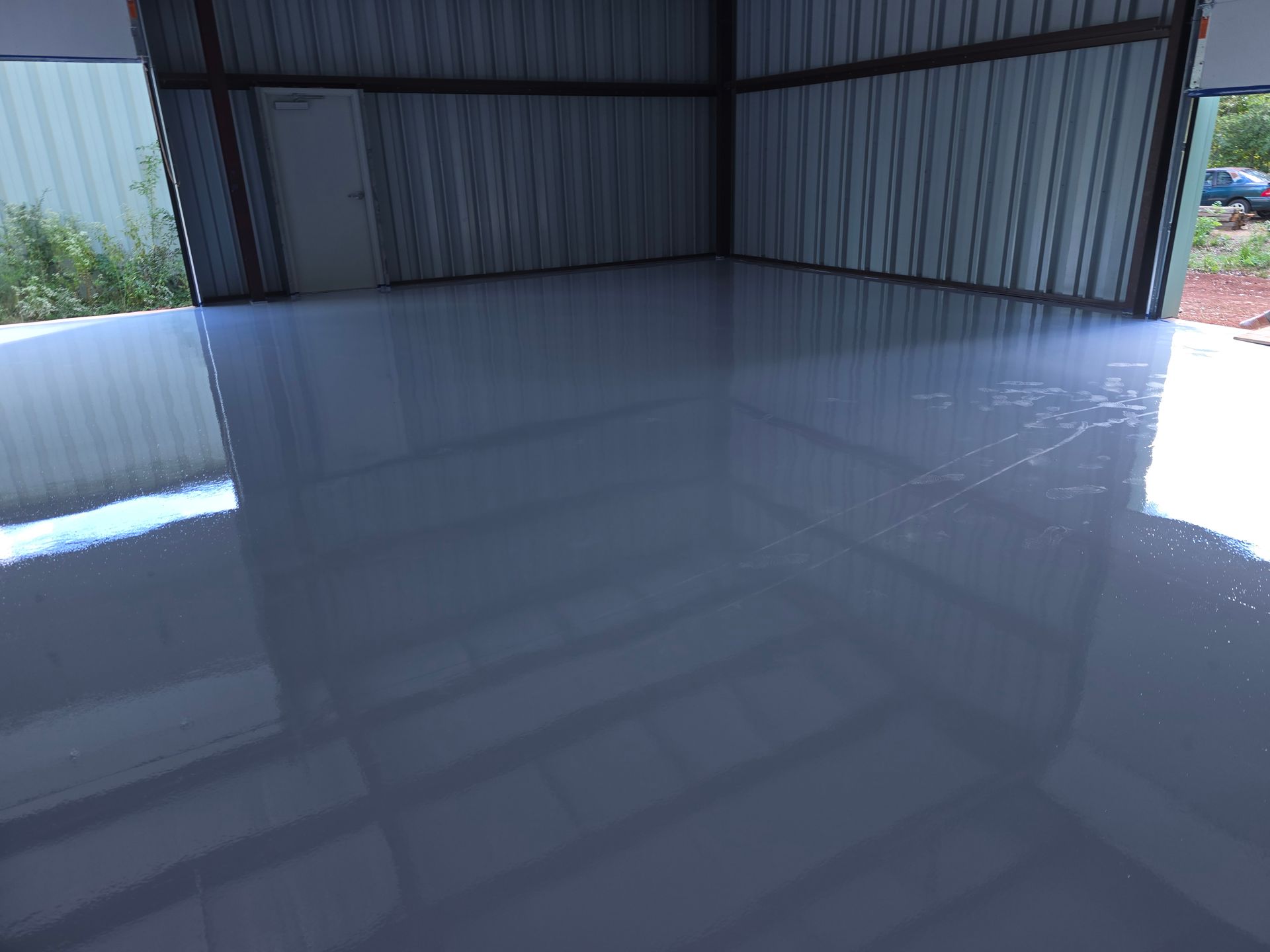 A shiny, light gray epoxy floor inside a metal-walled building, with a door and open entrance visible.