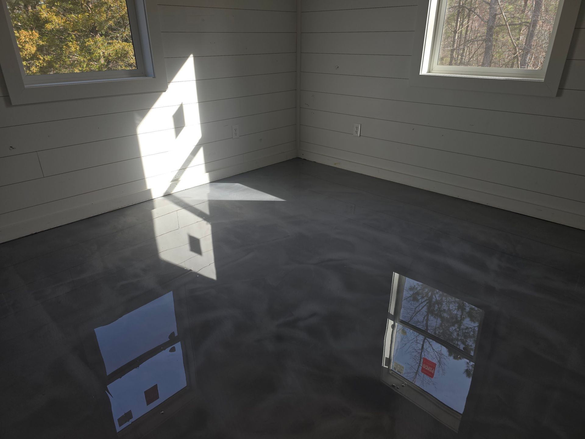 A room with a glossy, gray floor reflects sunlight from two windows. The walls are white with horizontal paneling.