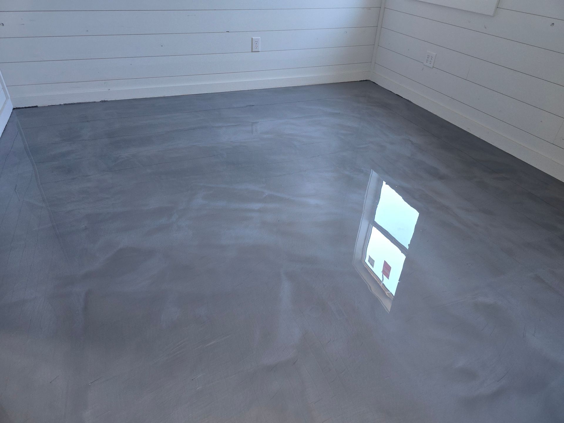 Gray, glossy epoxy floor reflecting light from a window in a room with white horizontal shiplap walls.