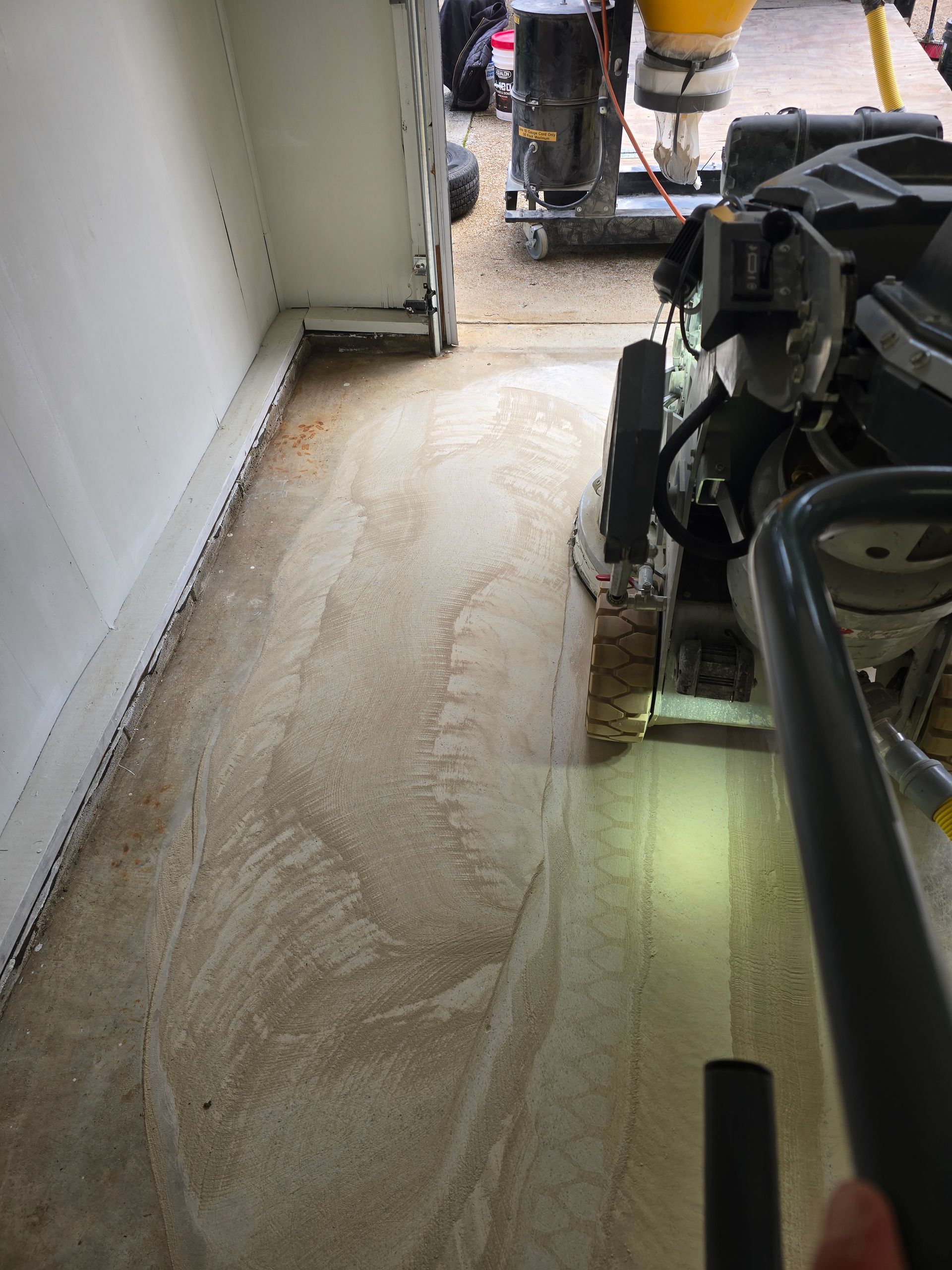 A concrete floor being ground with a power tool. The floor is light colored with visible swirls and the tool is black.