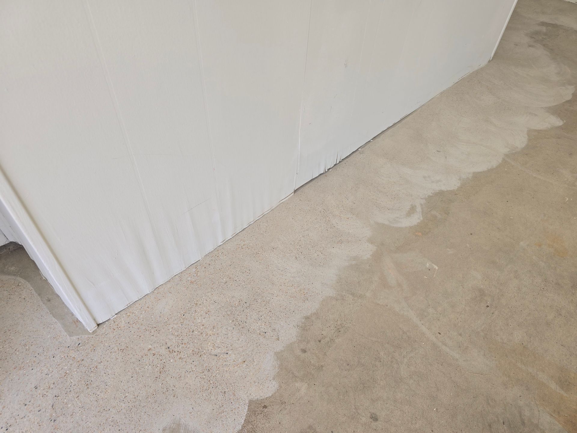Water damage on a concrete floor, spreading from a white wall's base. Pale water stains the floor.