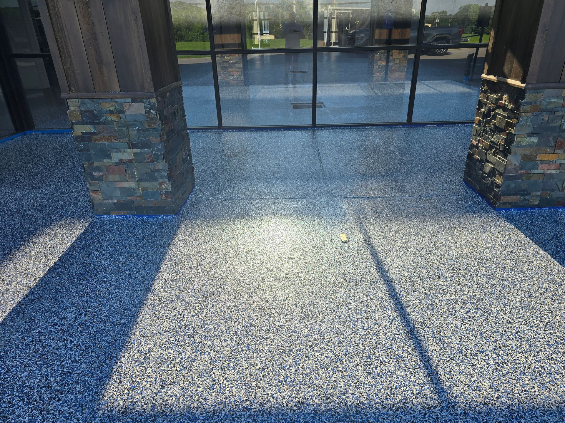 Blue and gray speckled flooring outside a building with glass windows and stone pillars; sunlight casts shadows.