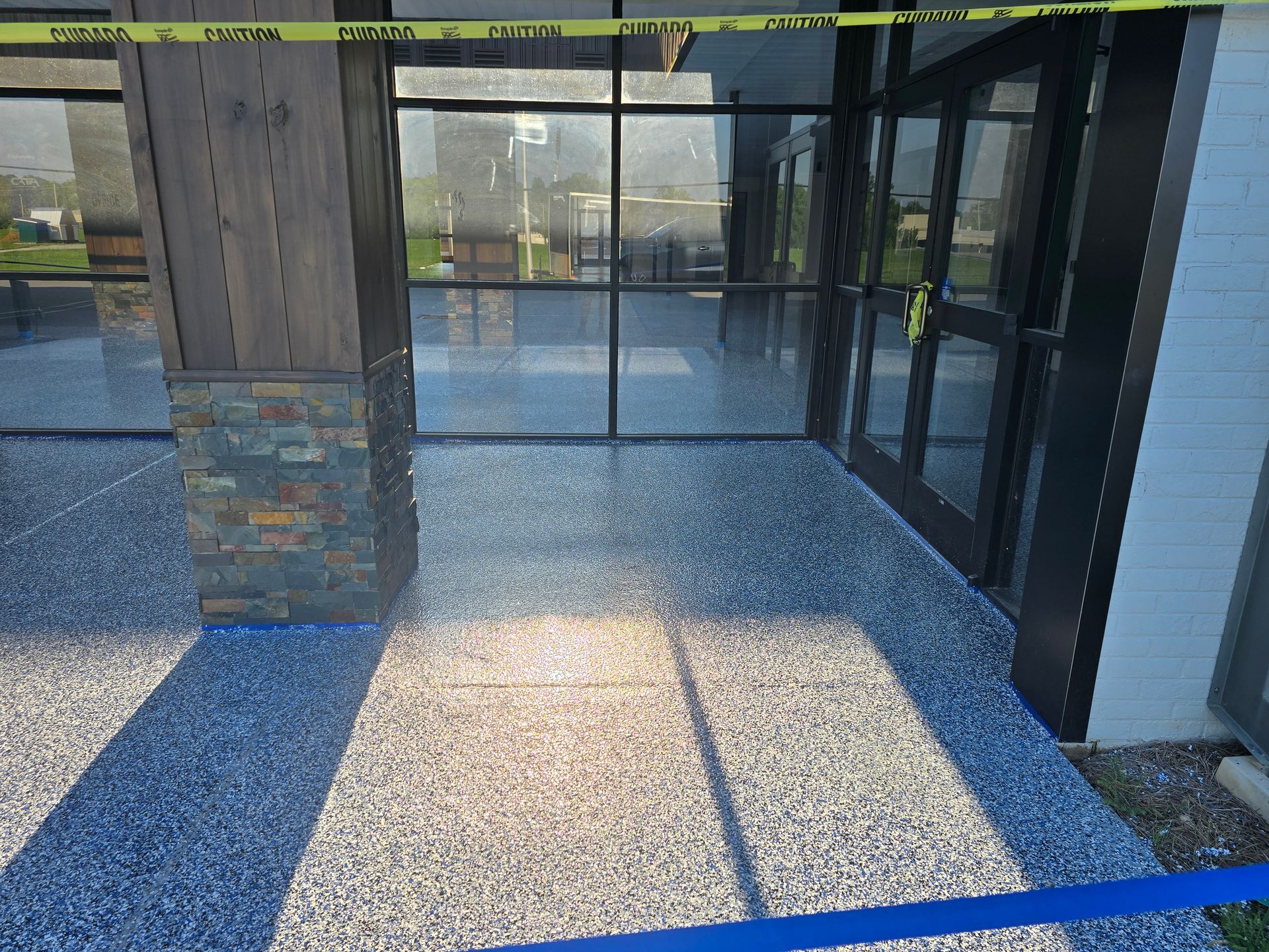 Entrance with a speckled gray floor and glass doors; a stone pillar is on the left. Yellow caution tape and a blue tape barrier are visible.