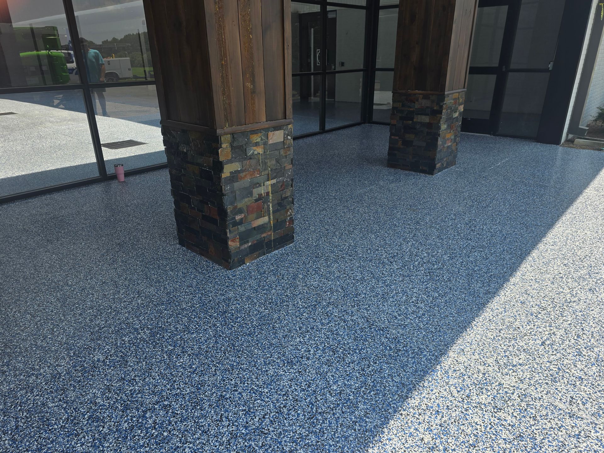 Exterior view of a building entrance featuring a speckled blue and white epoxy floor and dark stone pillars. Glass doors and windows visible.