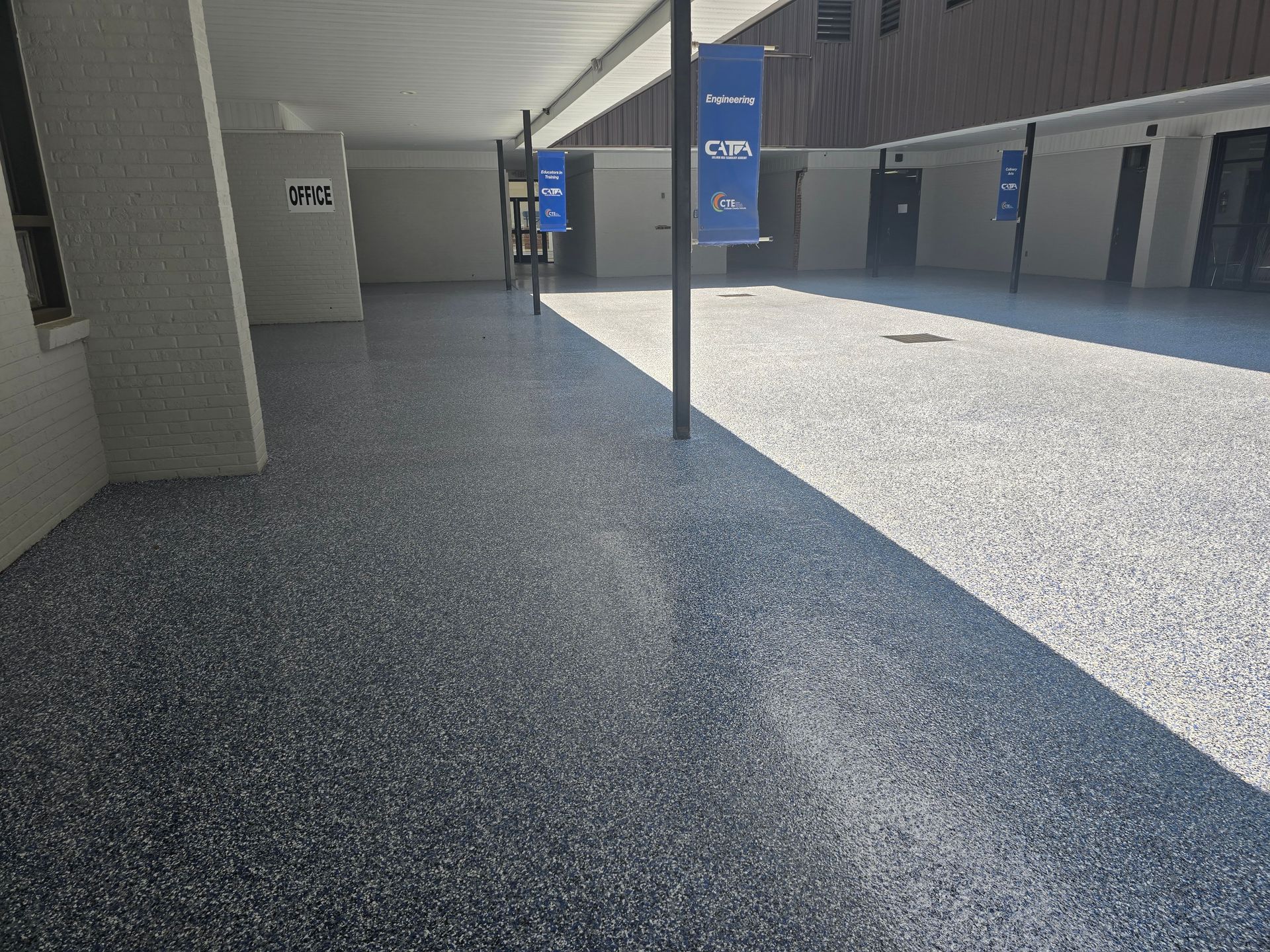 Covered walkway with a speckled, polished floor. Sunlight casts a long shadow across the floor. Banners hang on the pillars in the background.