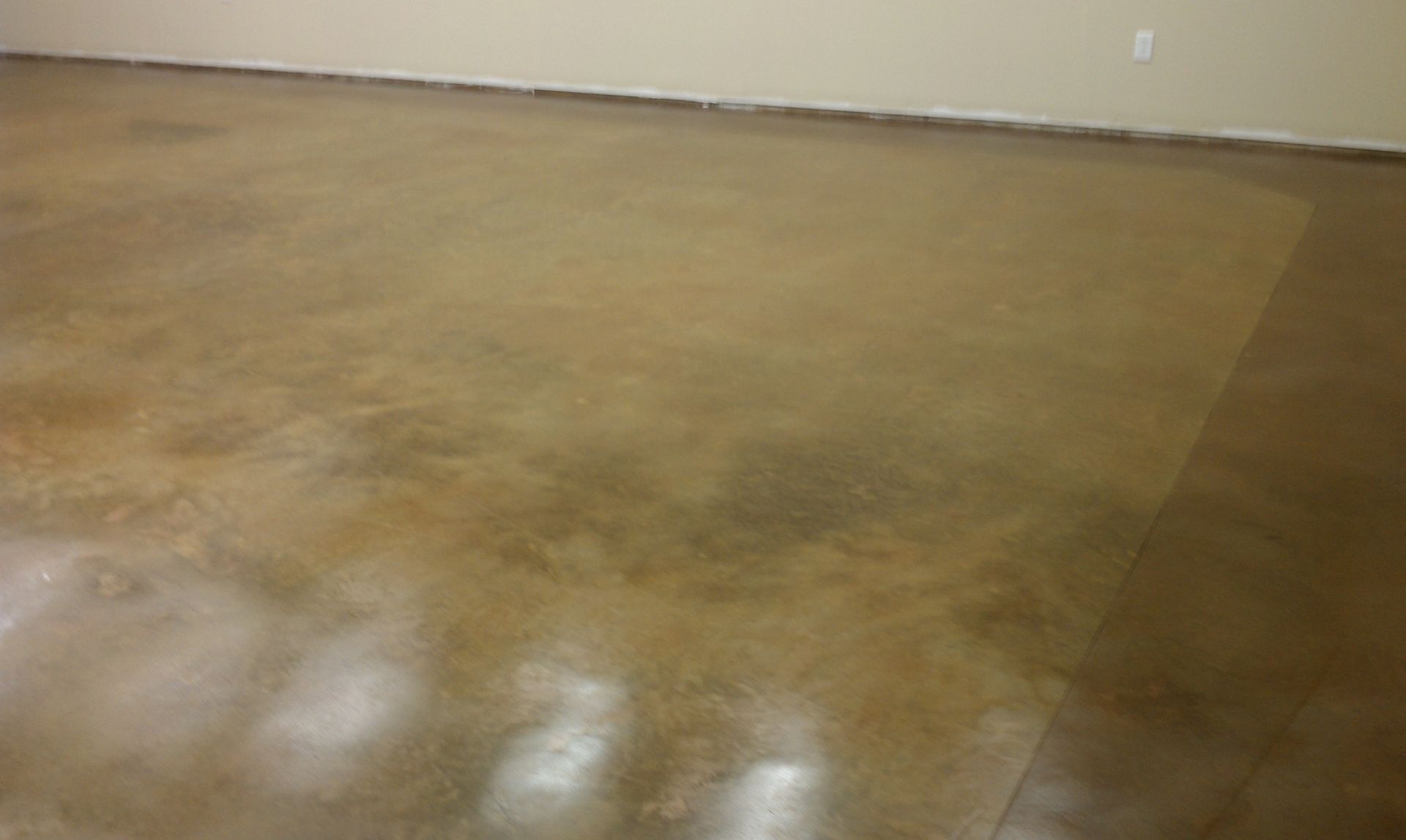 Brown stained concrete floor in a room, with lighter and darker areas. The floor meets a white wall at the top.