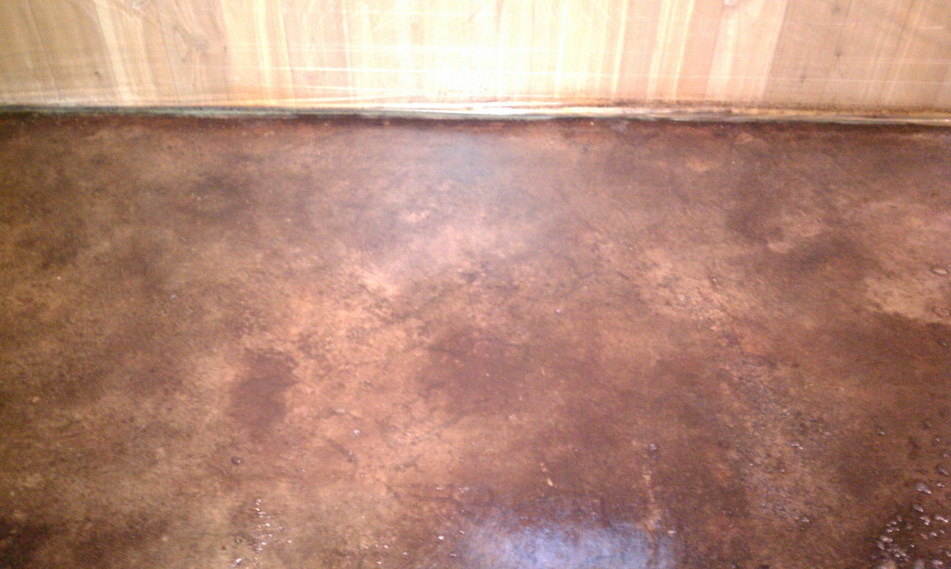 A close up of a stained brown concrete floor with a wooden wall in the background.