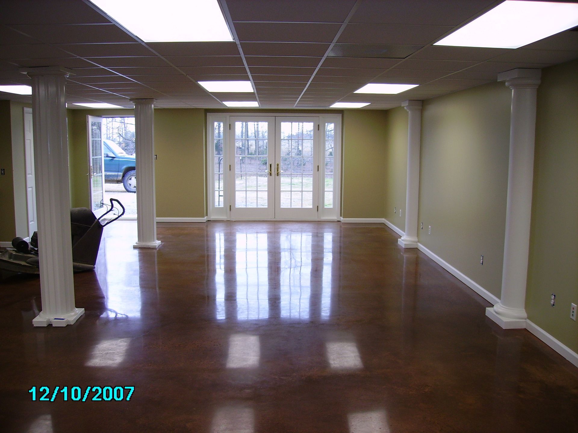 Spacious basement room with a polished brown floor, white pillars, and French doors leading outside.